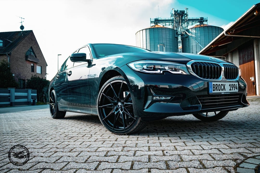 Black BMW sedan with custom wheels parked on cobblestones against a background of metal silos and blue sky.