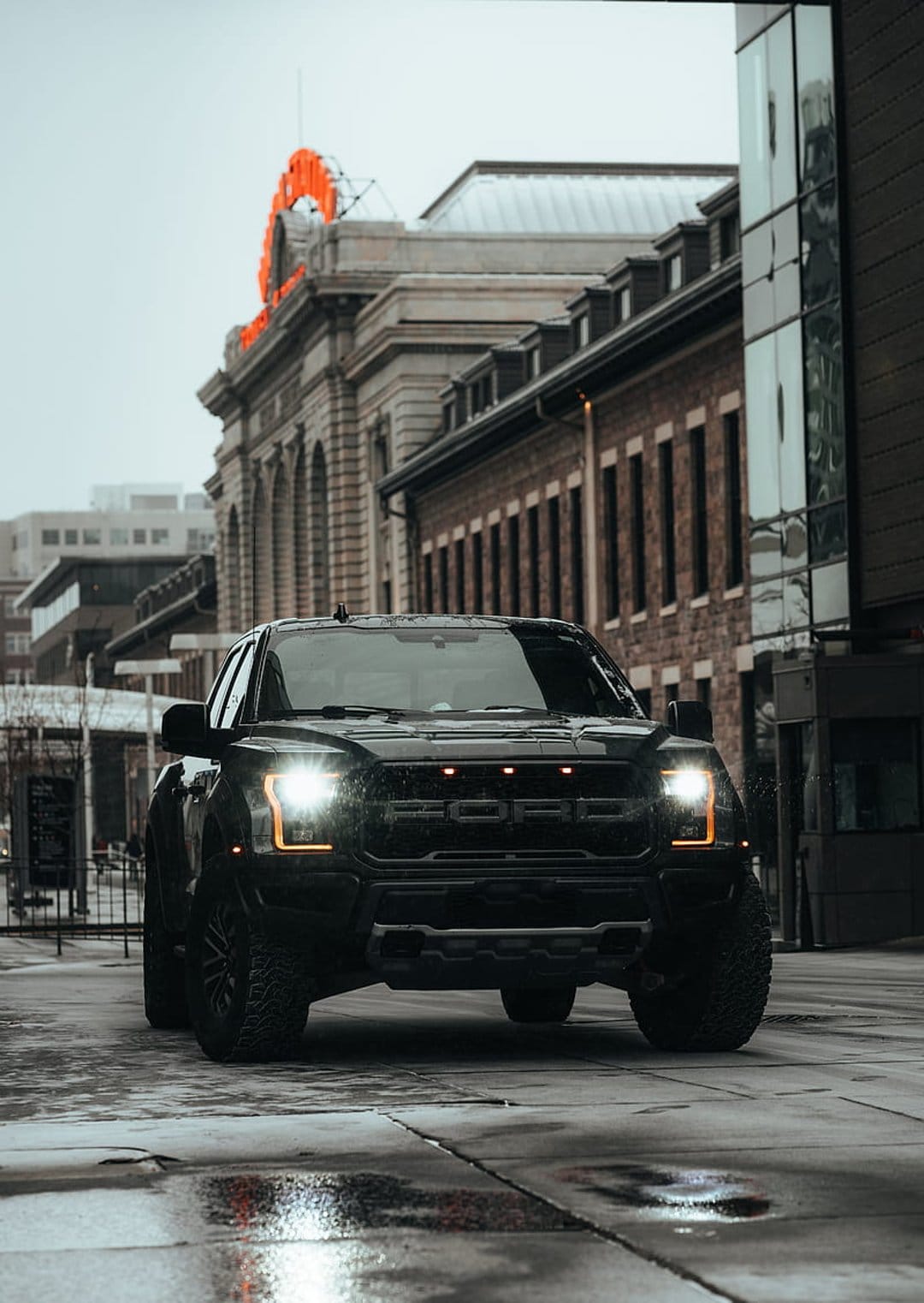 Black Ford F-150 Raptor truck with glowing headlights parked on a wet street reflecting city buildings.