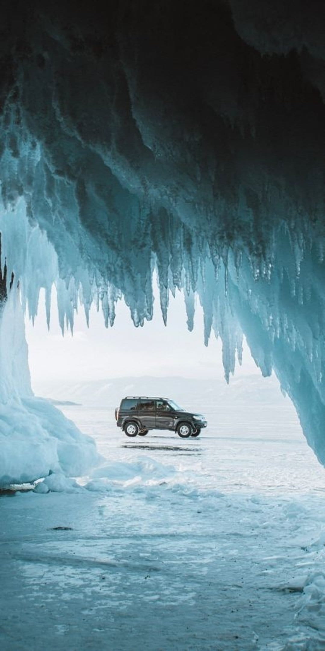 Dark SUV driving on a frozen lake seen through the opening of an ice cave with hanging icicles during winter.