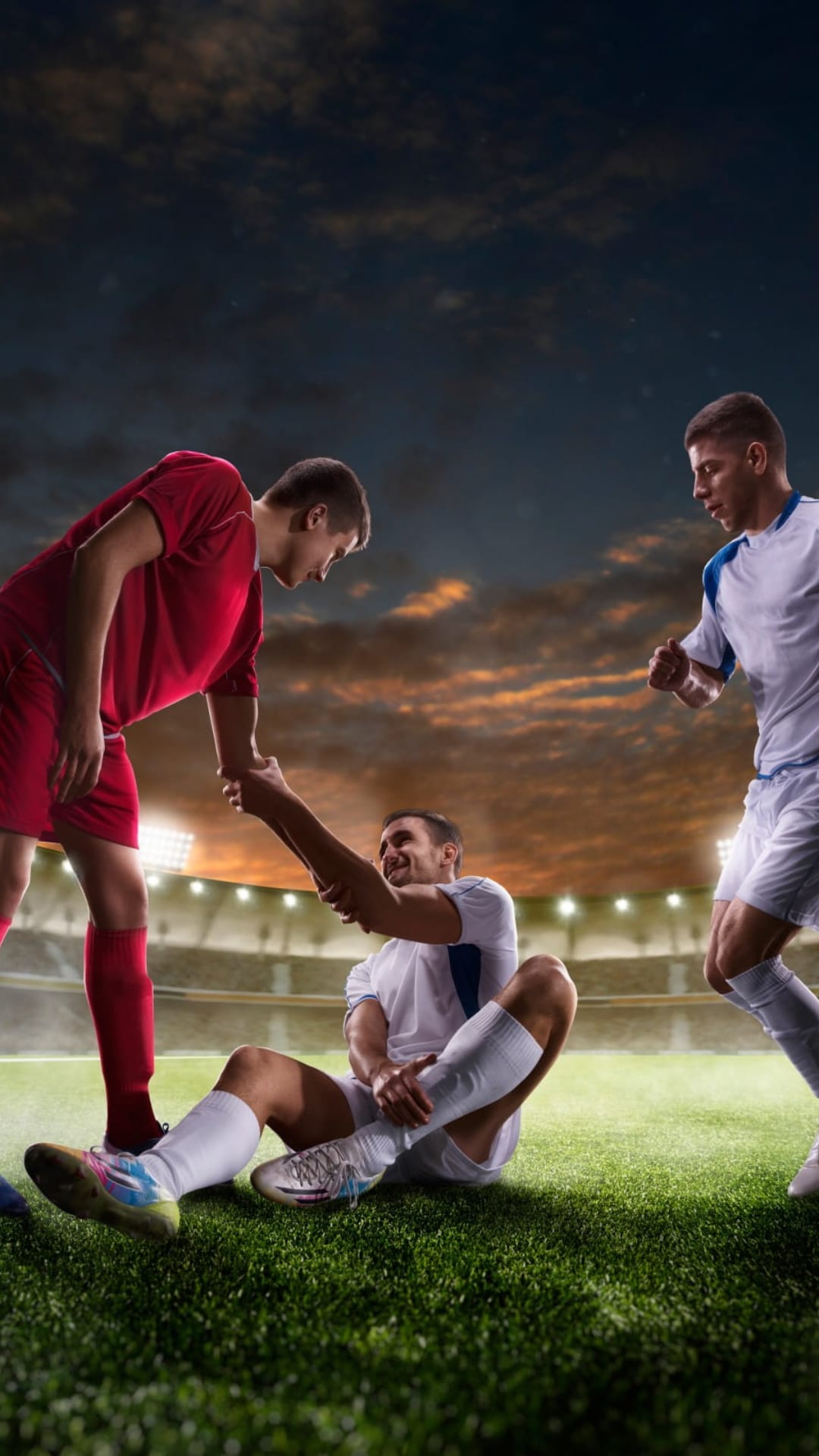 Soccer player in red extends a hand to help an injured athlete on a grass field under bright stadium lights at dusk.