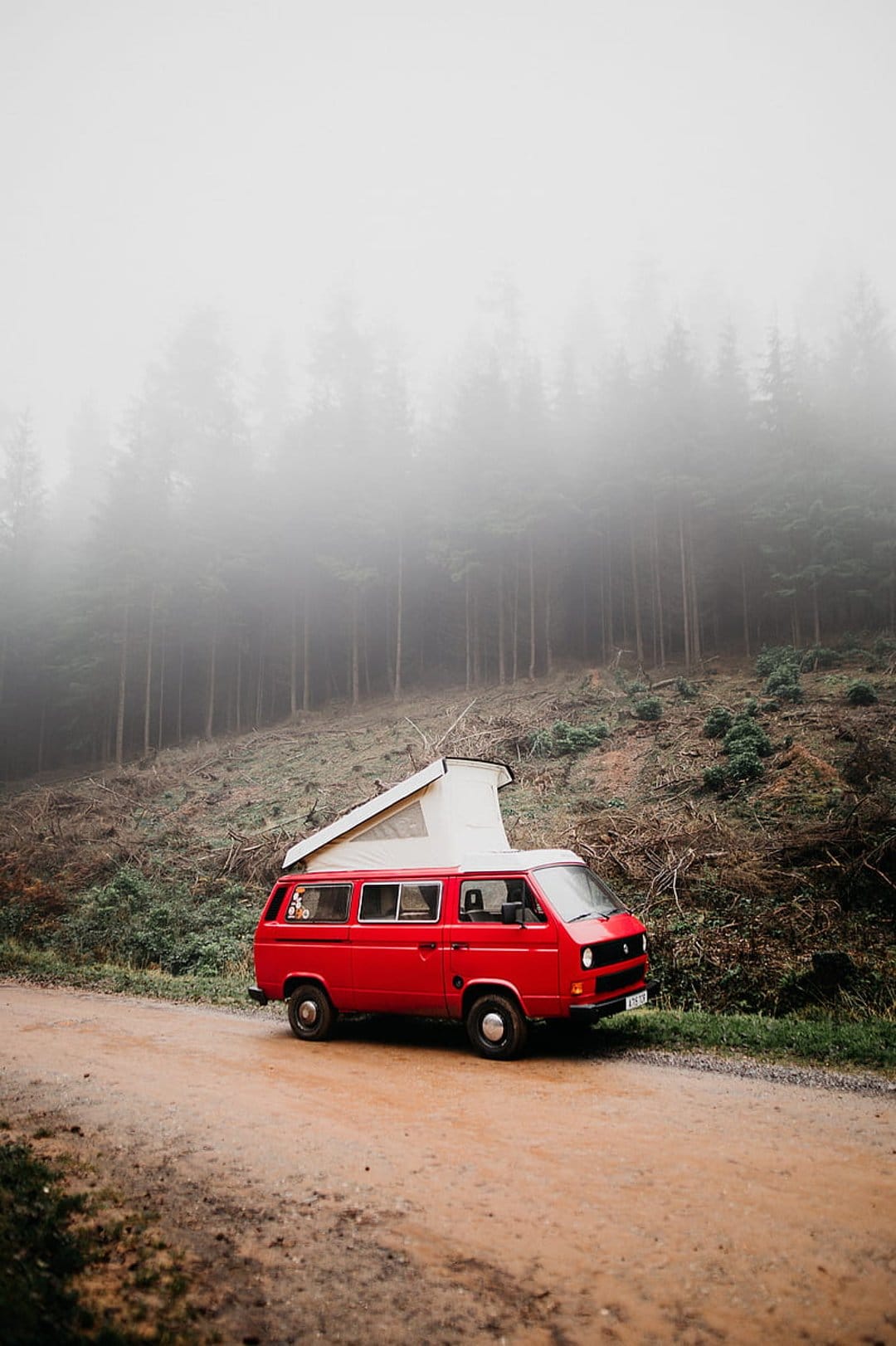 Vintage red camper van with a pop-top roof parked on a muddy forest road under thick, atmospheric fog.