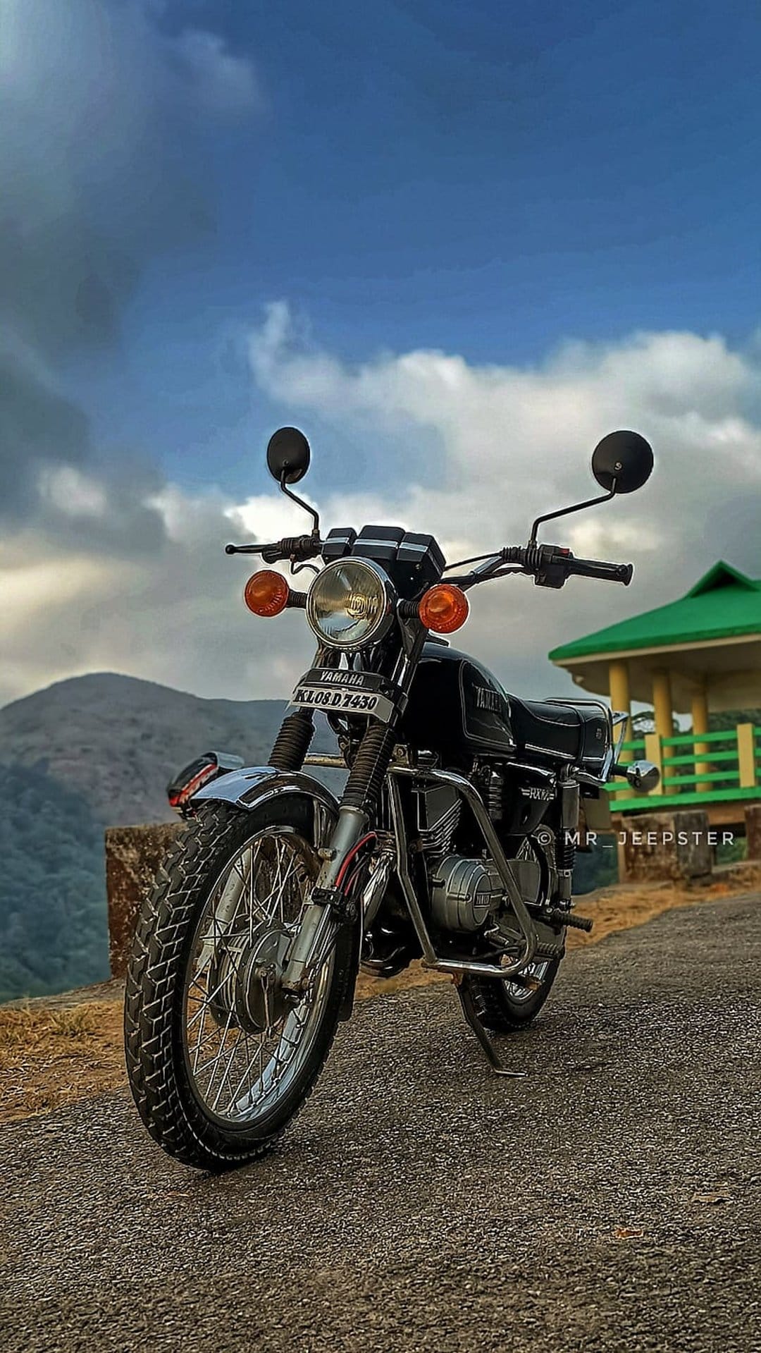 Black Yamaha RX 100 vintage motorcycle on a gravel road with misty green mountains under a cloudy gray sky.