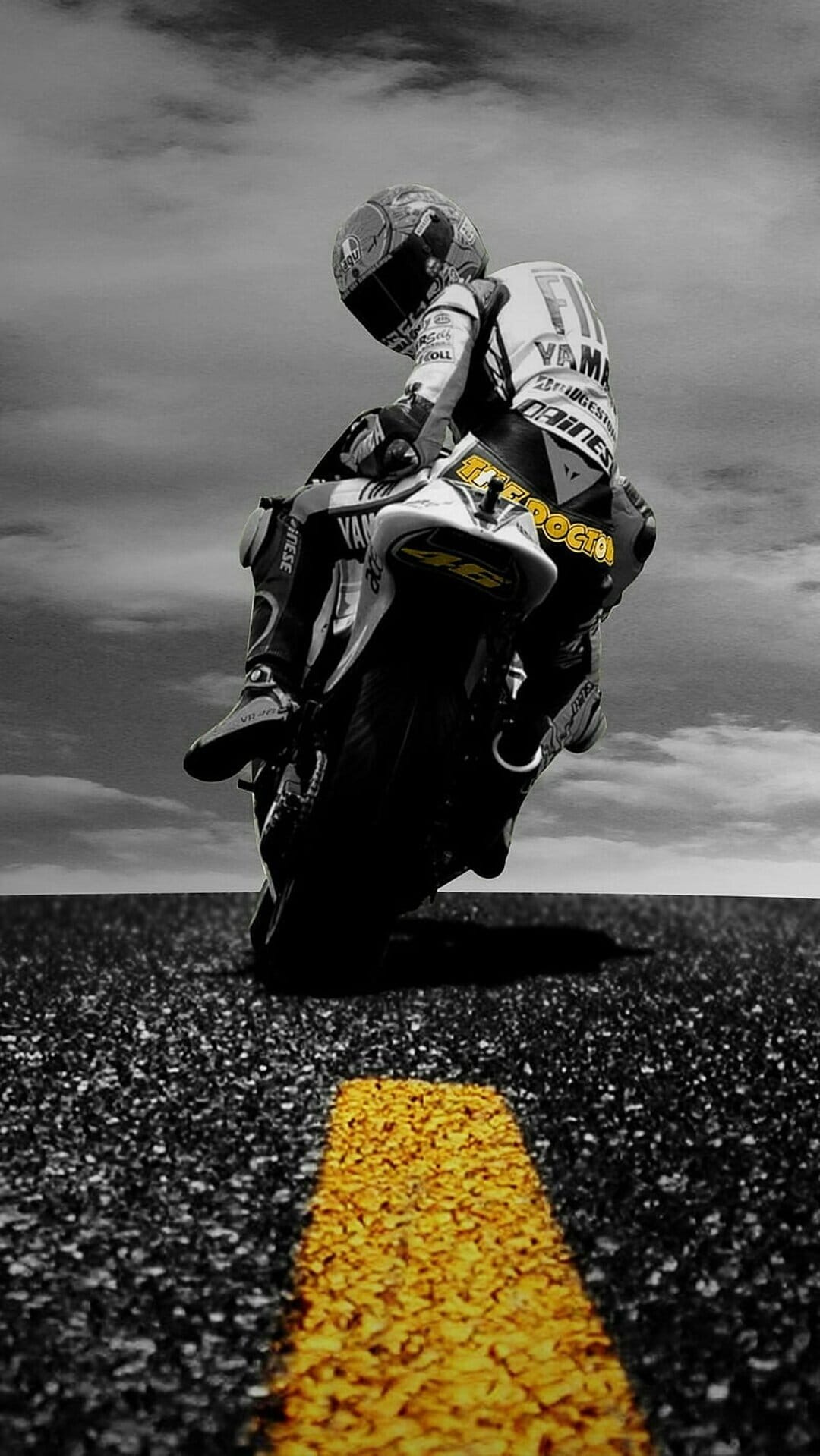 Motorcycle racer leans low on dark asphalt with a yellow stripe against a cloudy monochromatic sky.
