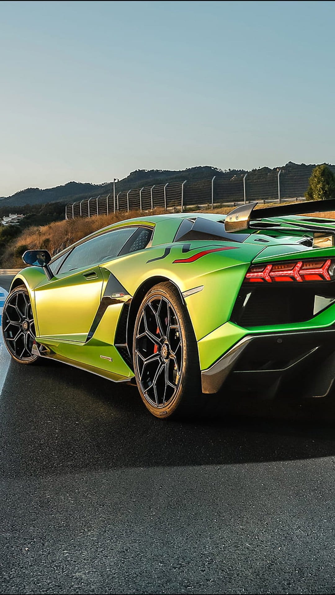 Green Lamborghini Aventador SVJ supercar on an asphalt track with black wheels and a large rear wing under a blue sky.