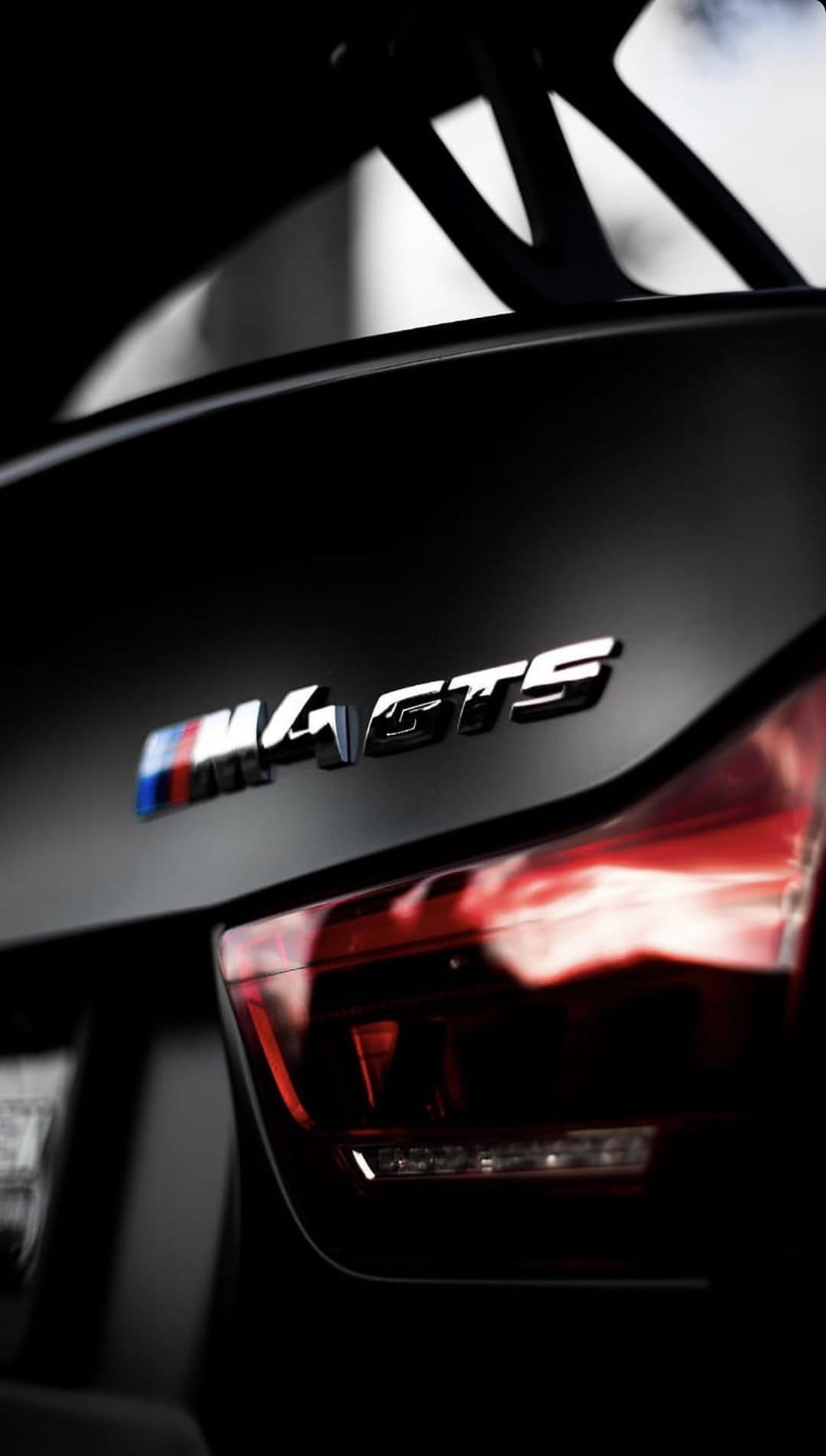 BMW M4 GTS rear emblem and glowing red taillights set against a dark matte black finish on a sleek car body.