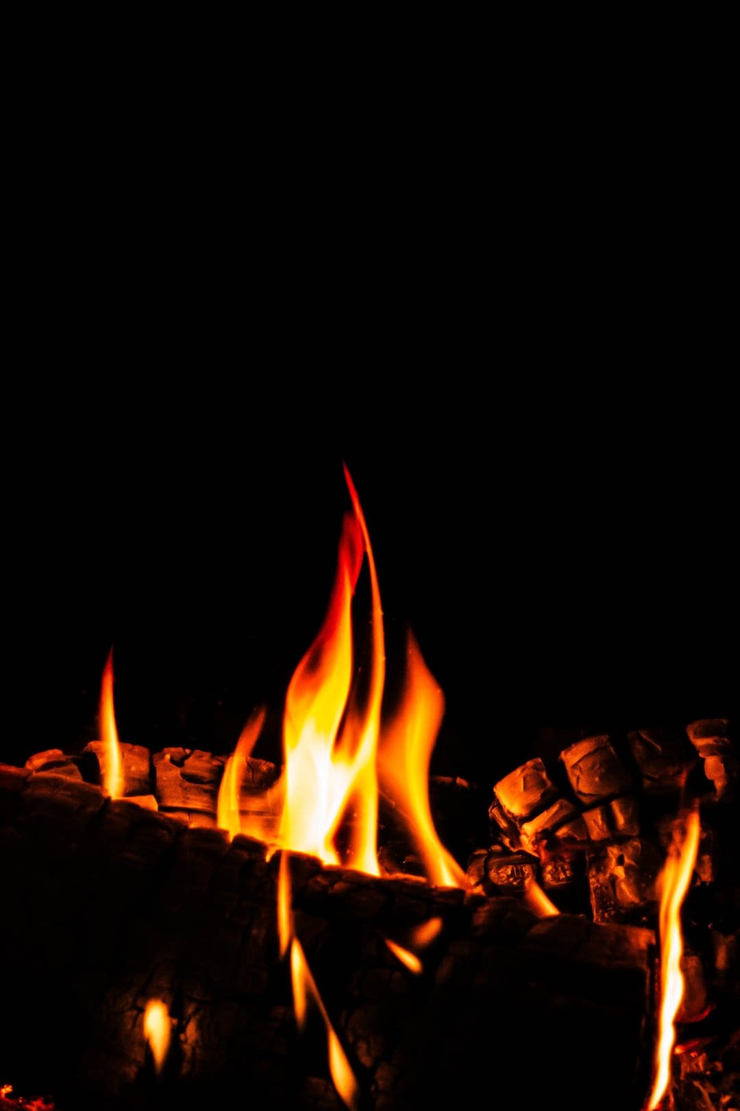 Vibrant orange flames and glowing embers rise from charred logs in a dark fireplace with soft lighting.