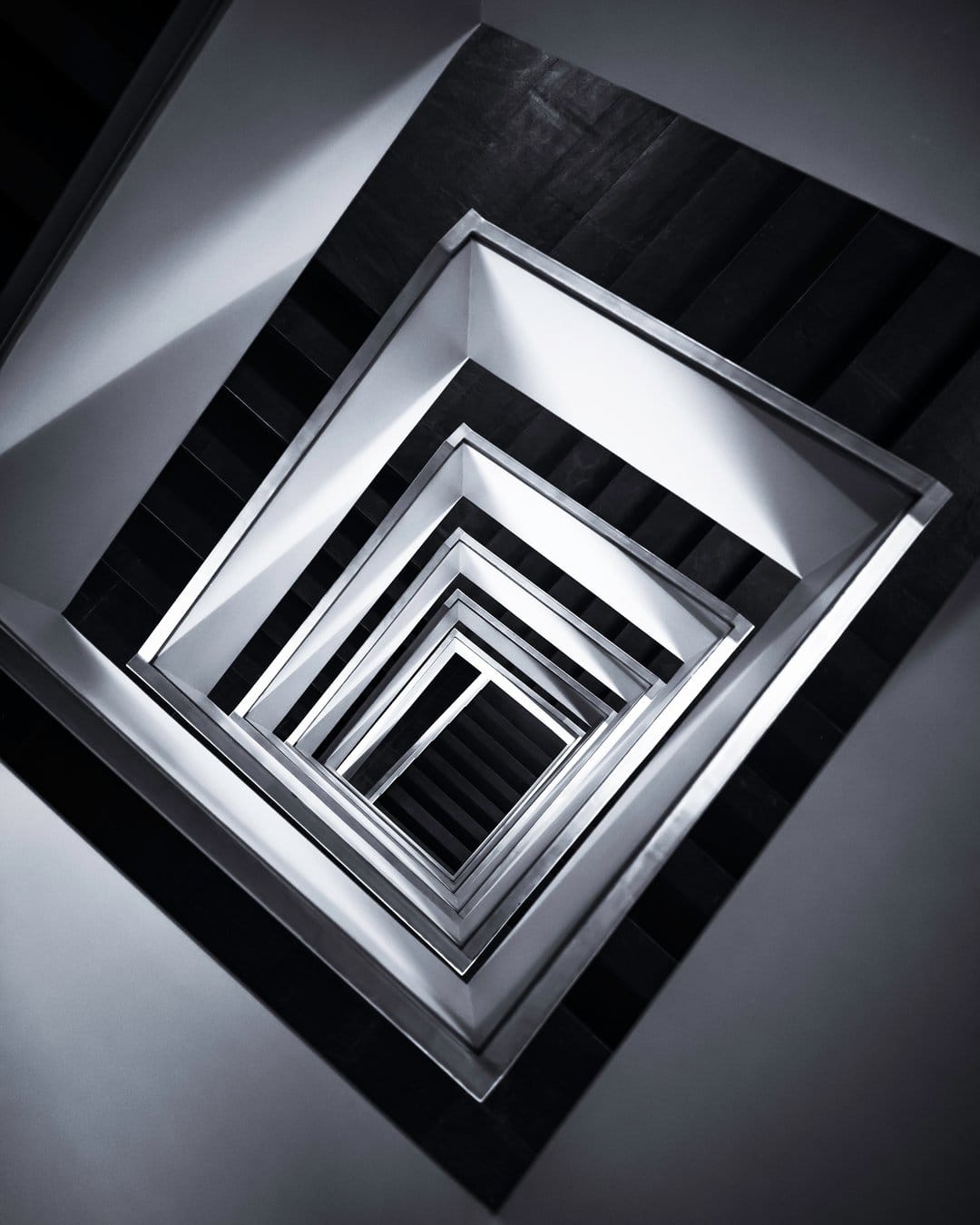 Square metallic railings form a receding geometric pattern with harsh light and dark shadows in a deep stairwell.