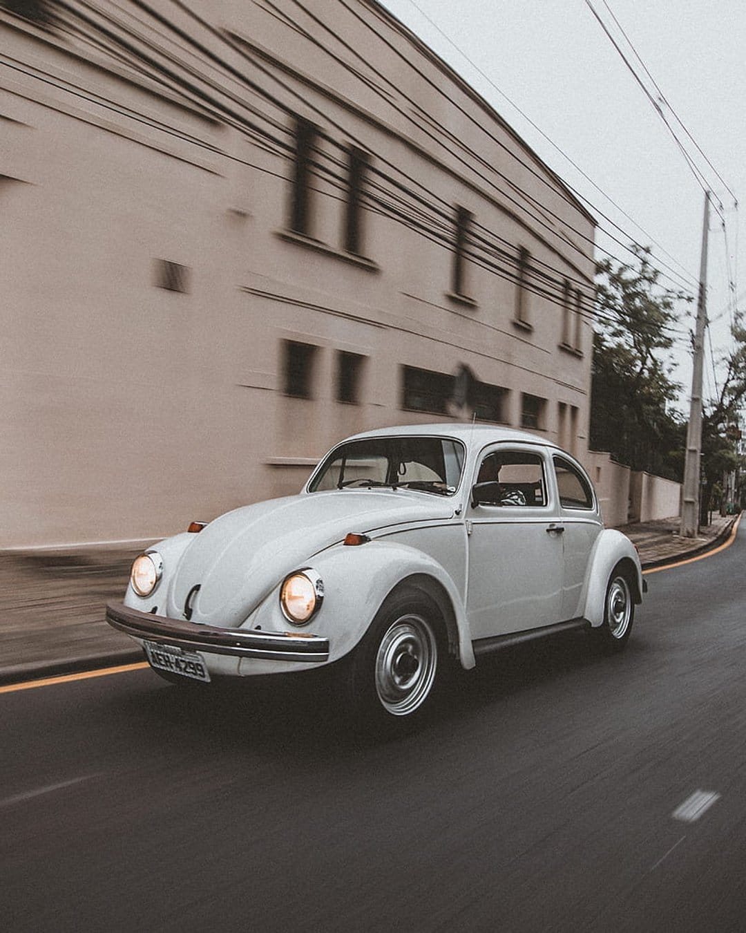 White Volkswagen Beetle with lit headlights drives down a tree-lined street with motion blur at sunset.