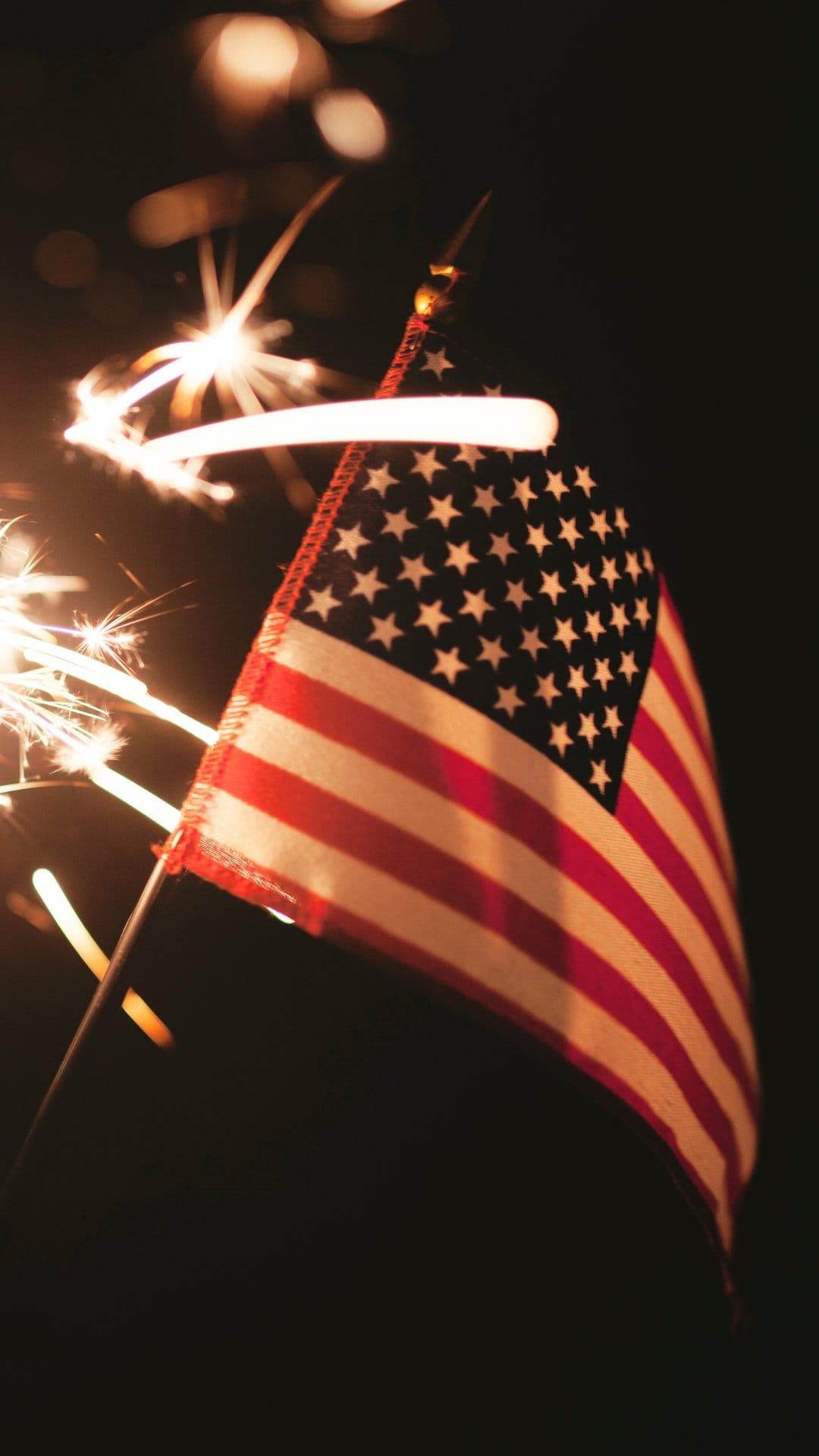 American flag ripples in the dark while illuminated by golden sparks and glowing light trails from a sparkler.