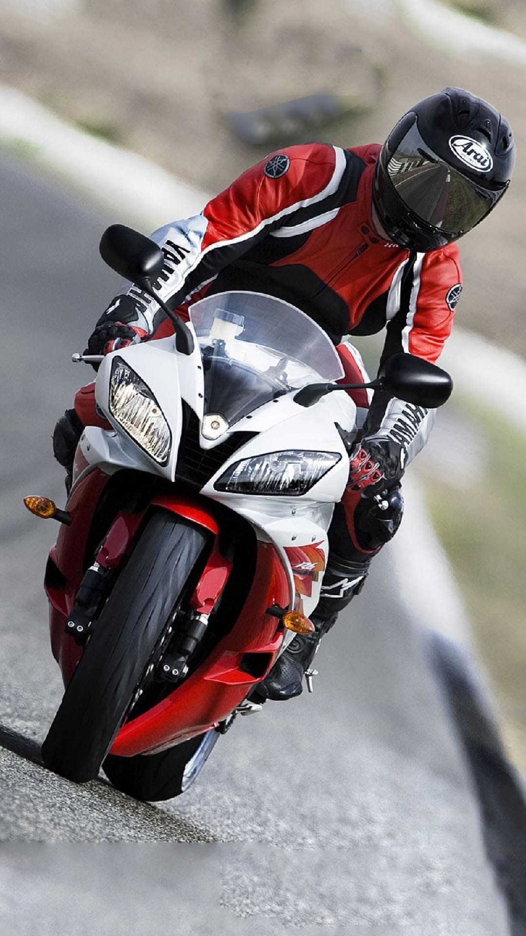 Motorcyclist in a red racing suit leans a sportbike deep into a turn on a textured asphalt racetrack.