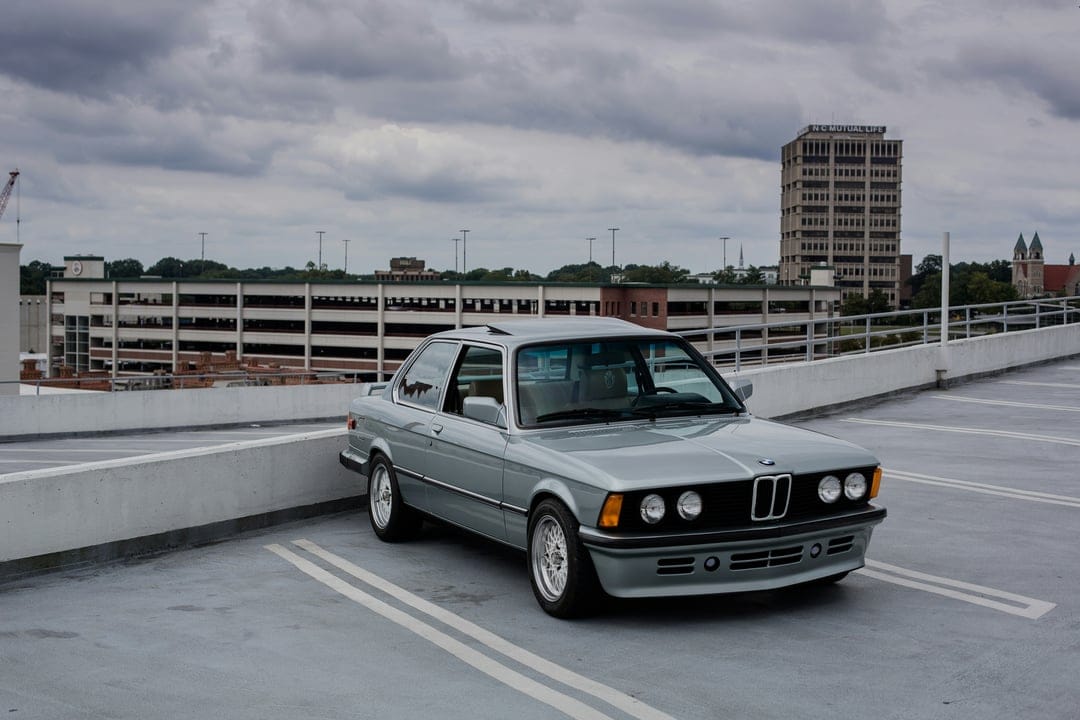 Silver BMW E30 coupe parked in a lot with a concrete barrier and a multi-story garage under an overcast sky.