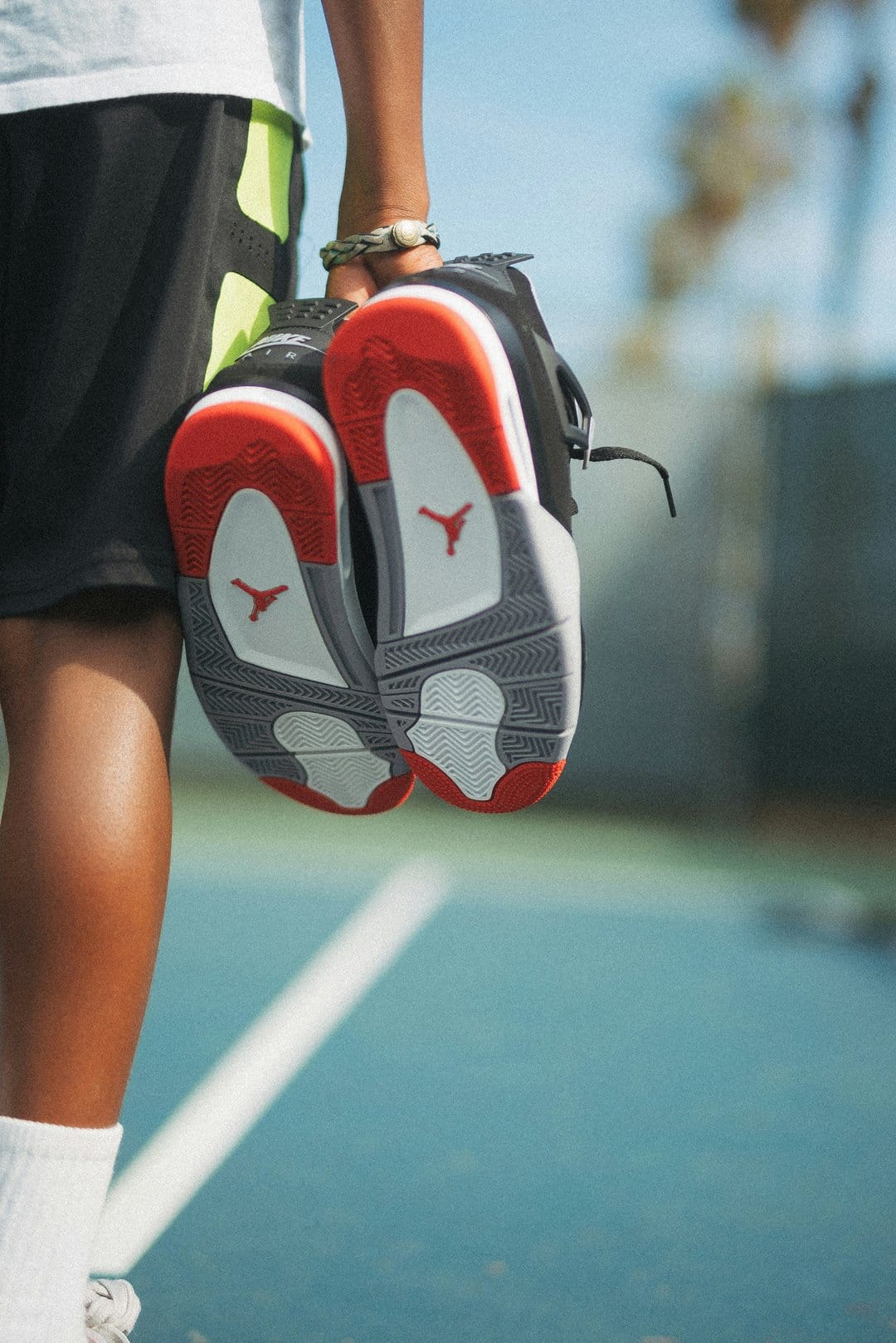 Person holding red and black Jordan sneakers over a blue tennis court, revealing the Jumpman logo on the soles.