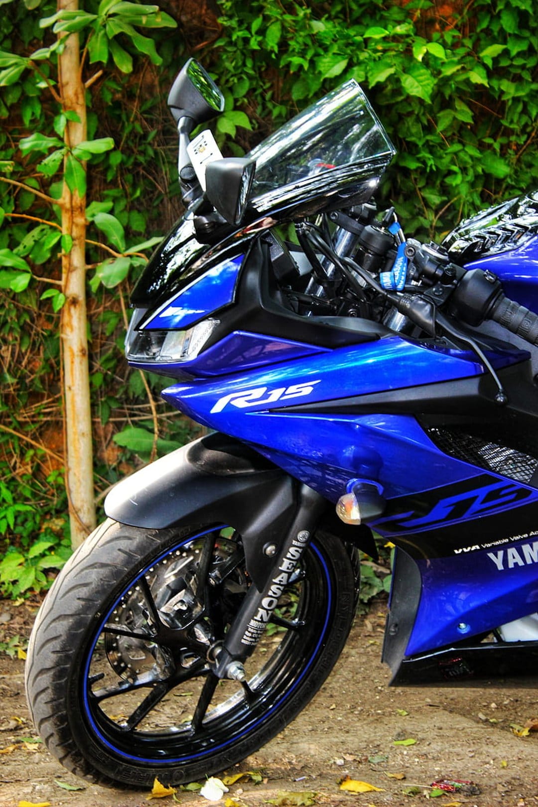 Blue Yamaha R15 motorcycle front wheel and fairing against blurred green trees in a vertical portrait view.