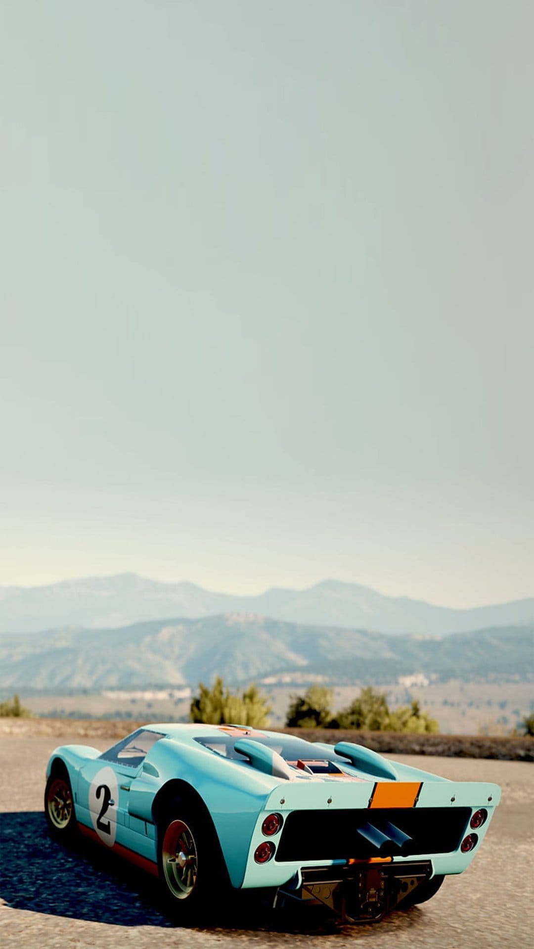 Blue Ford GT40 race car with orange stripes parked on a dusty mountain road overlooking a scenic valley vista.