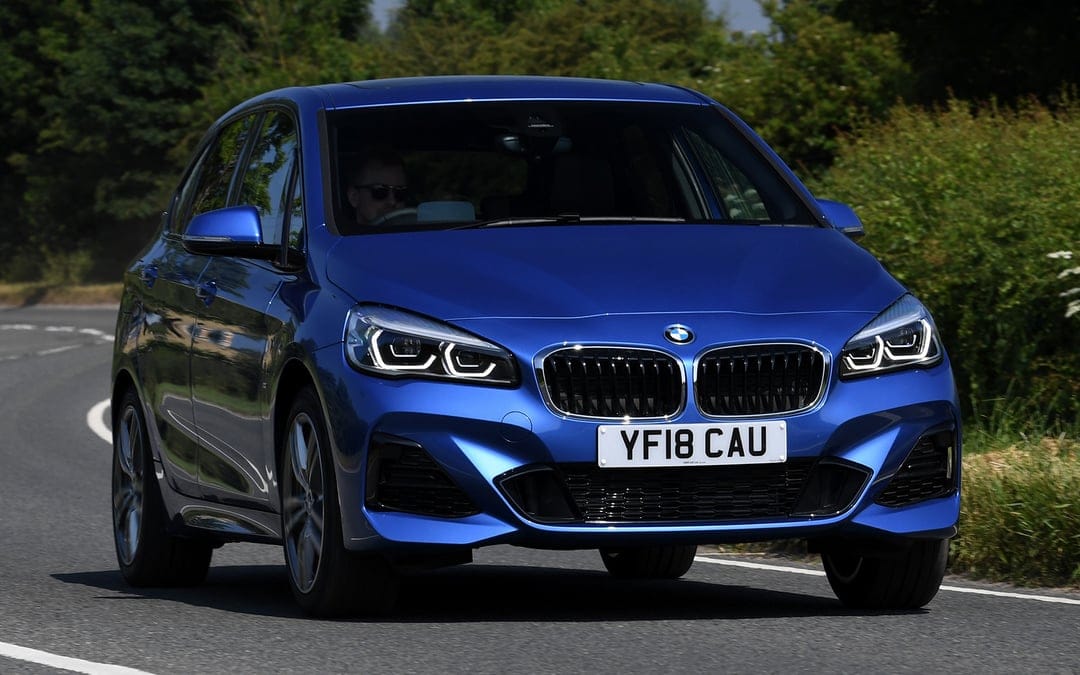 Blue BMW 2 Series car driving on a paved road surrounded by green trees under bright daytime sunlight.