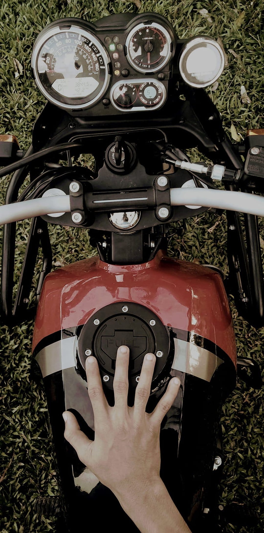 Hand resting on a black motorcycle fuel tank with a 'FUEL' cap and dashboard gauges over a grassy surface.