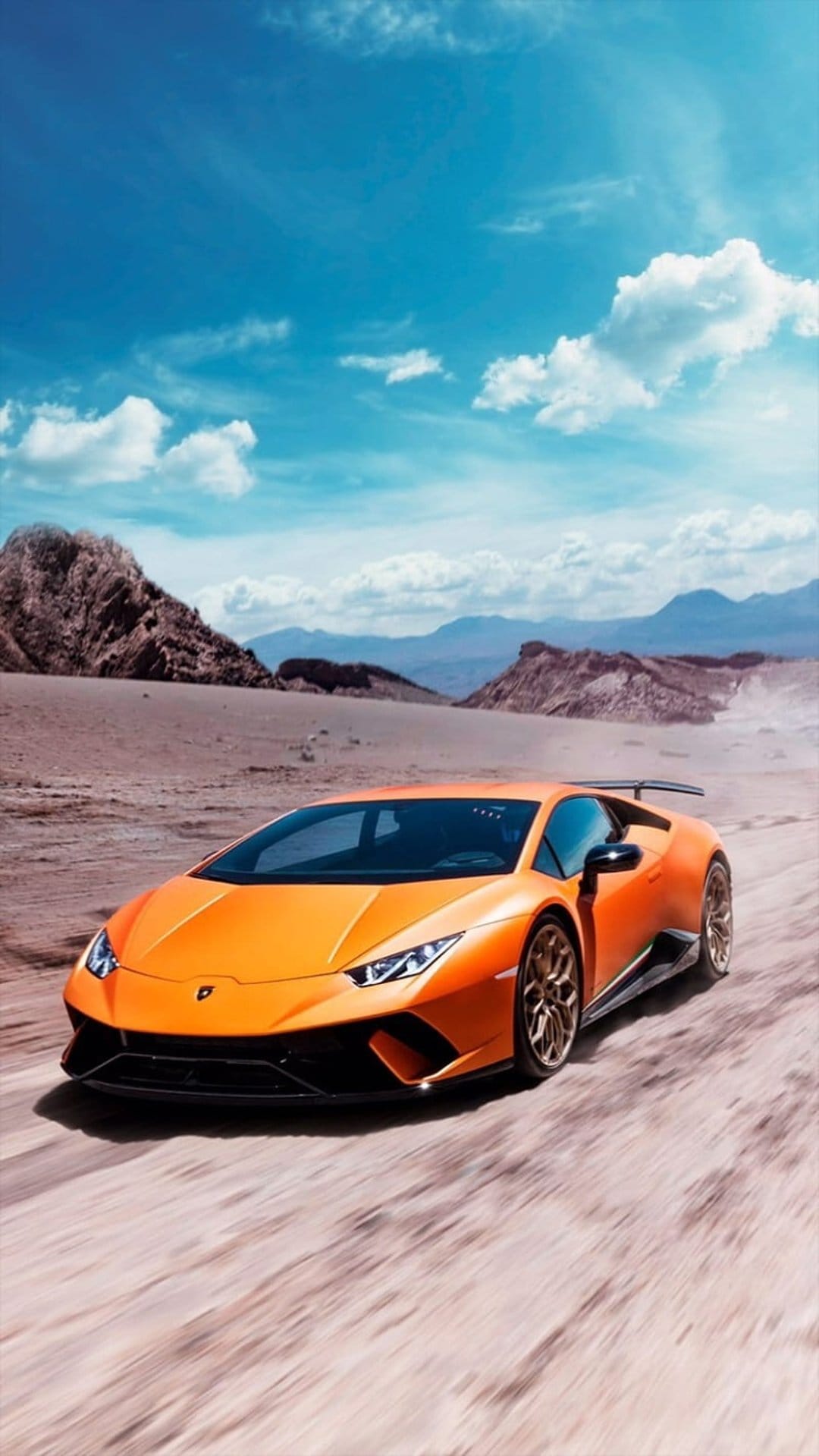 Orange Lamborghini supercar kicks up dust while racing through a sunny desert landscape under a blue cloud sky.