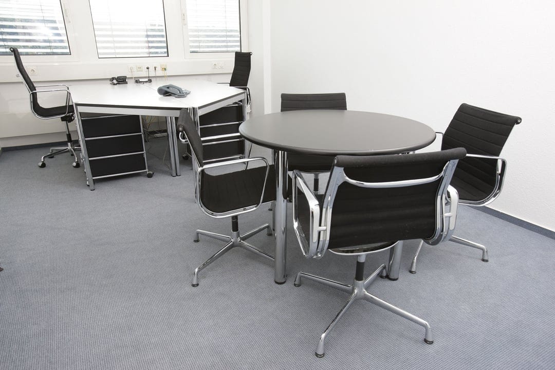 Black mesh executive chairs surround a circular conference table in a modern office with large window blinds.