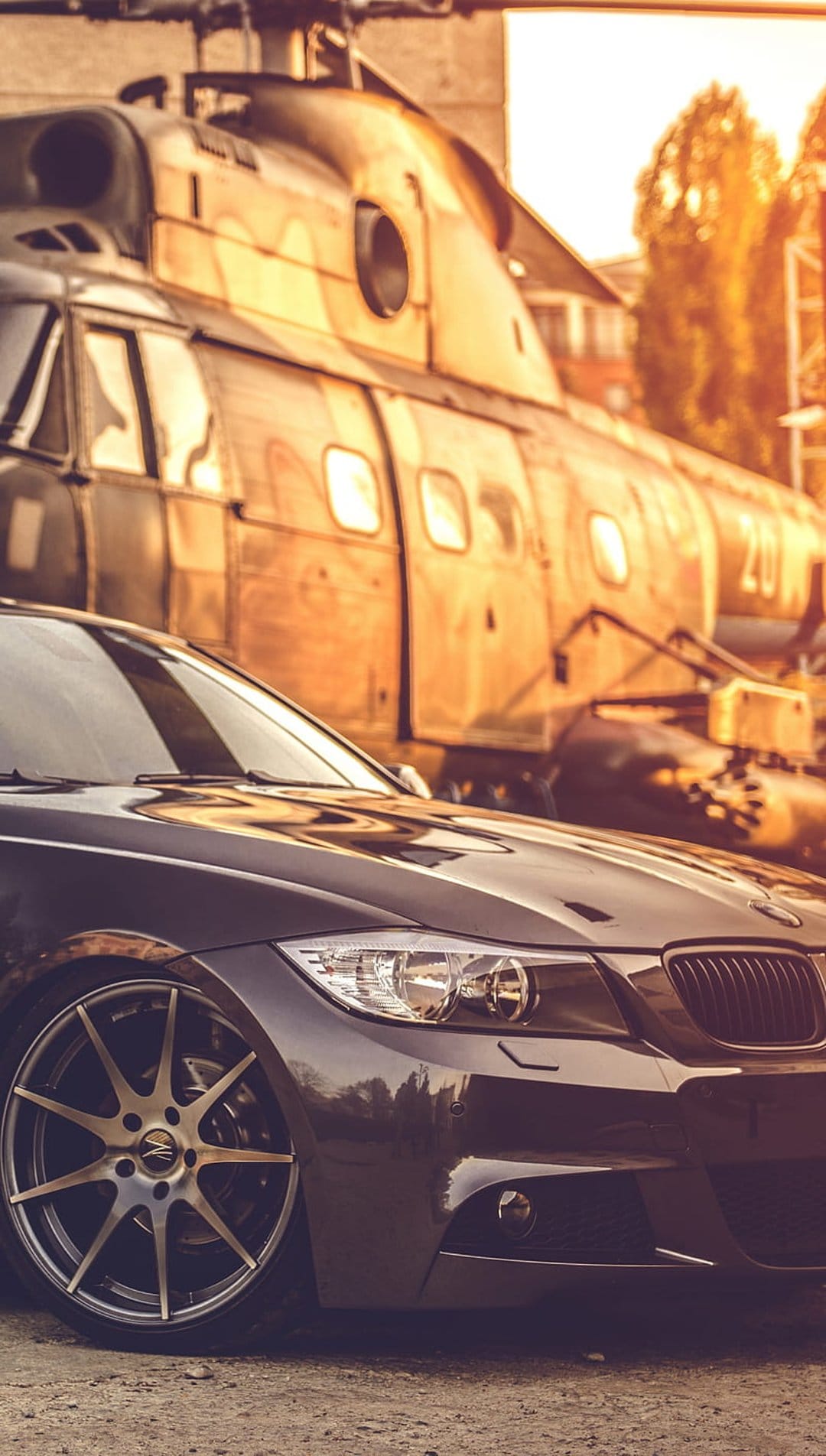Black luxury sedan parked beside a vintage helicopter during a vibrant golden hour sunset with warm light reflections.