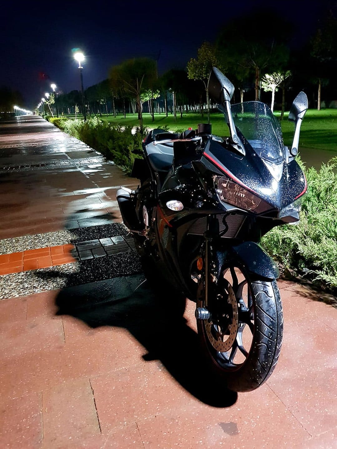 Black sport motorcycle parked on a wet stone path at night with streetlights reflecting in rain puddles.