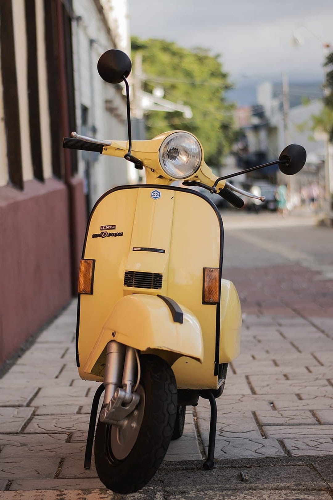Yellow vintage Vespa scooter parked on a gray cobblestone street with blurred buildings and greenery behind it.