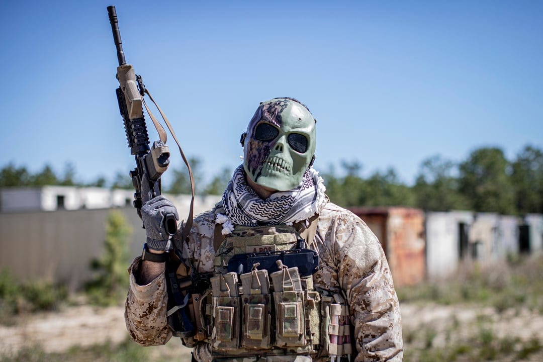 Soldier in a green skull mask and camouflage gear holds a rifle against a desert background with abandoned ruins.