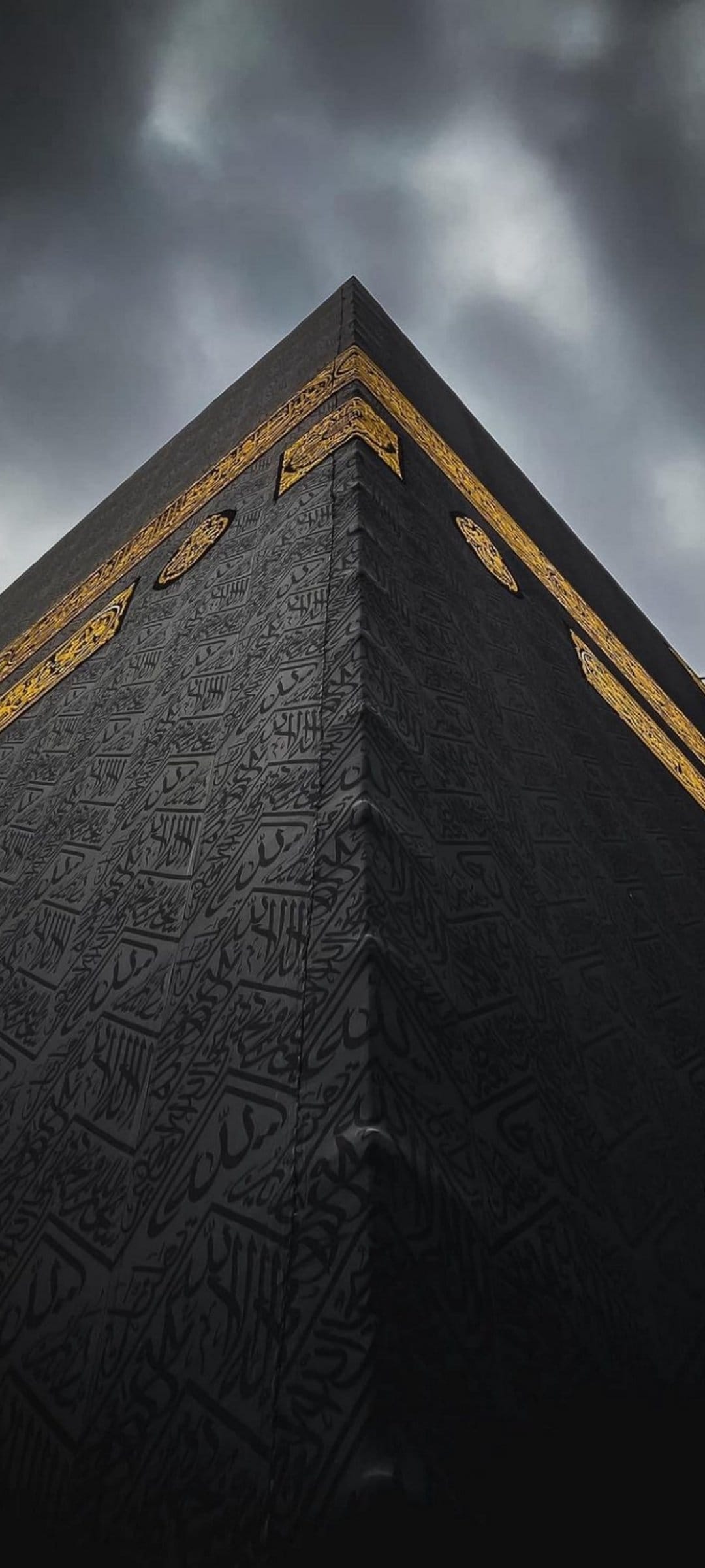 Kaaba corner draped in black silk with gold Arabic calligraphy against a dark, cloudy sky in a vertical view.