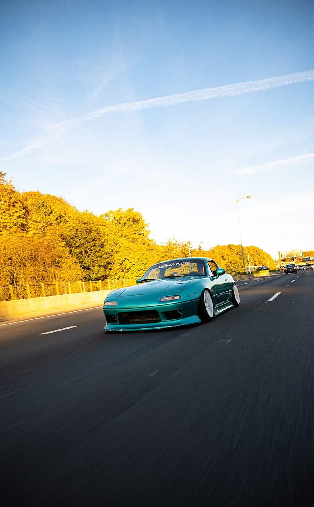 Teal Mazda Miata with white wheels driving on an autumn highway flanked by yellow trees under a blue sky.