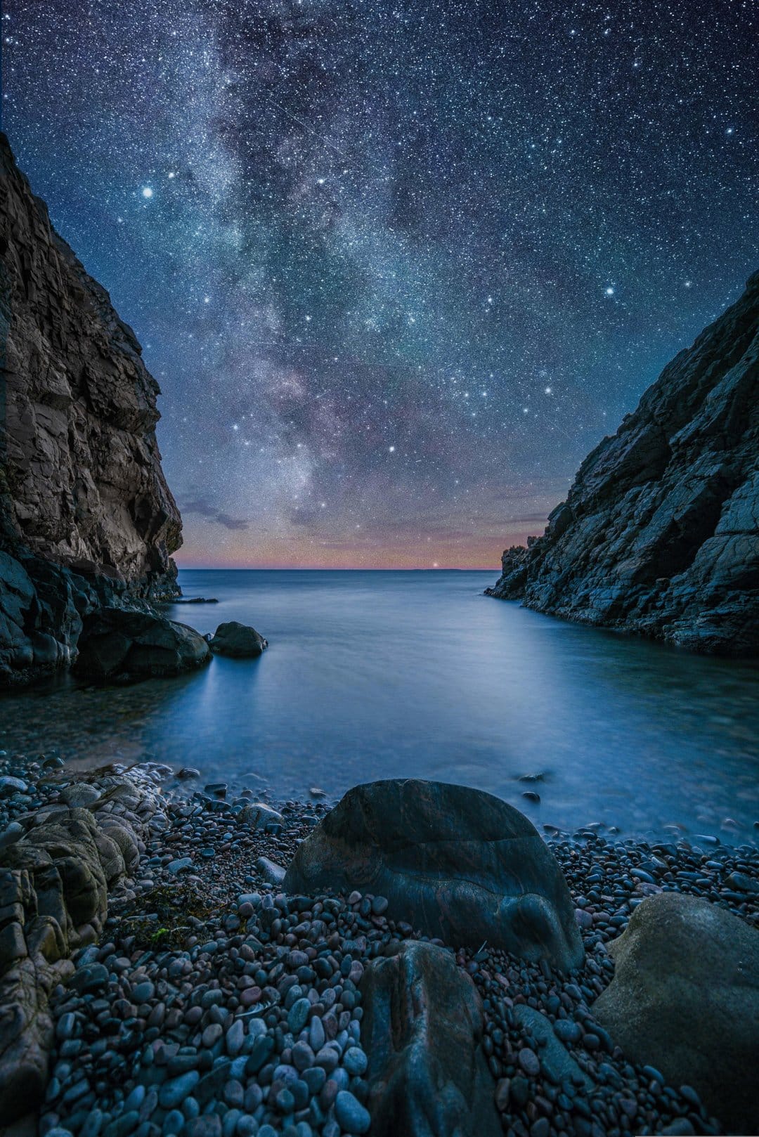 The Milky Way galaxy glows over a dark rocky cove and calm ocean water under a dense field of brilliant stars.