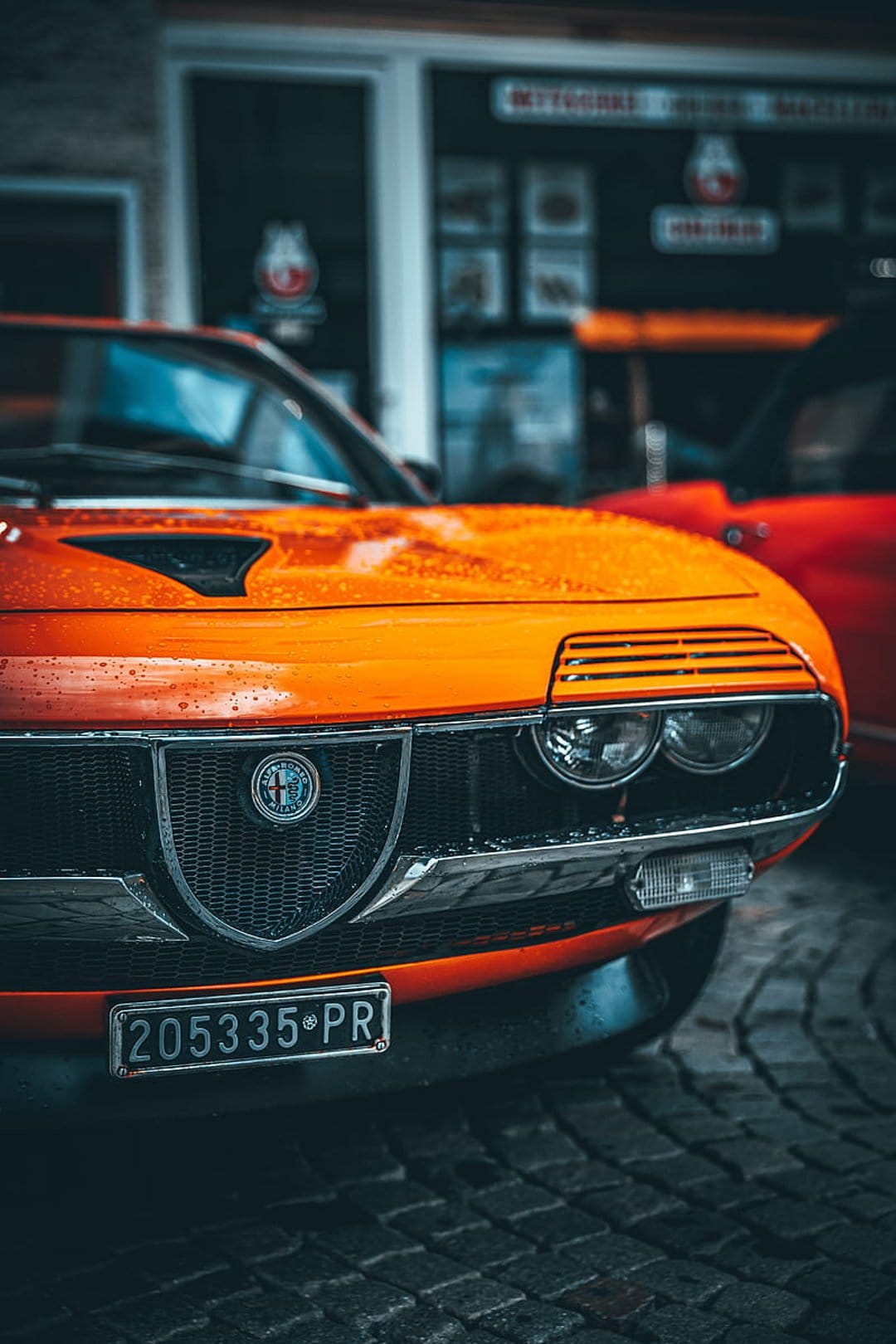 Vintage orange Alfa Romeo grille with rain droplets, round headlights, and a centered silver badge on cobblestones.