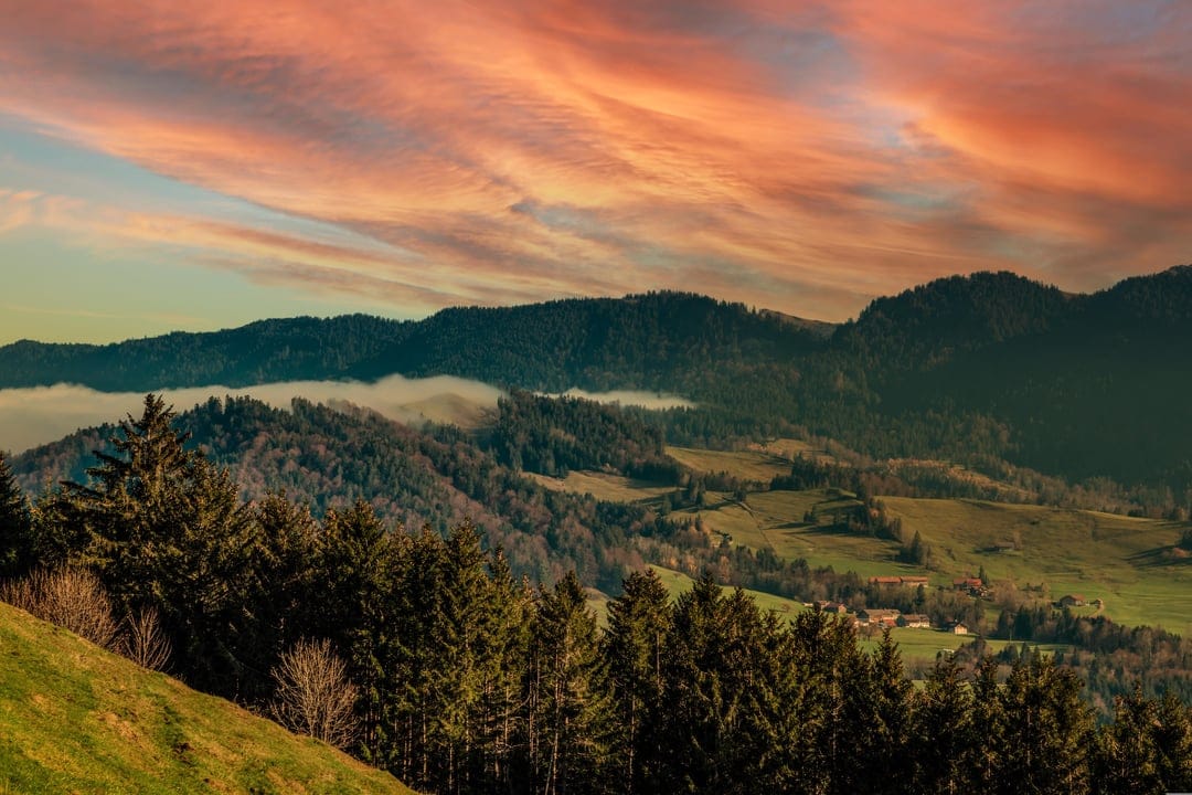 Rolling forested hills under a fiery orange and pink sunset sky with thin white mist settling in the deep valleys.