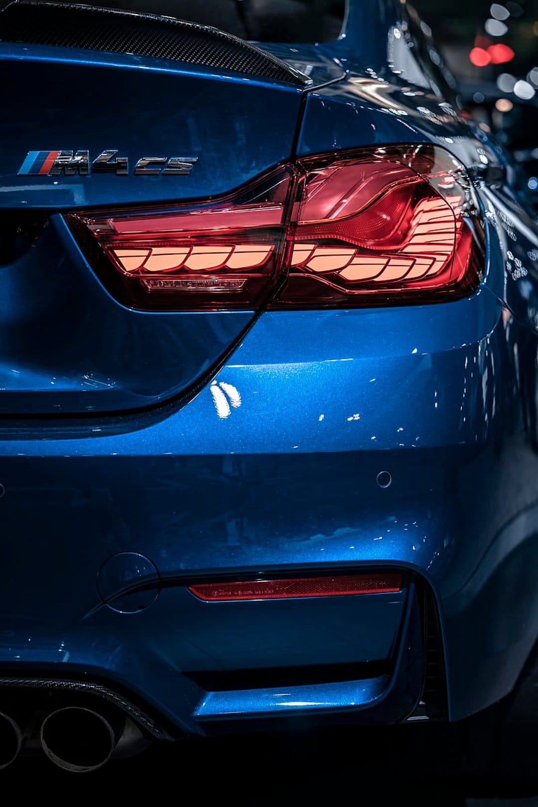 BMW M4 CS rear corner showing illuminated red LED taillights, blue metallic paint, and the M4 CS badge detail.