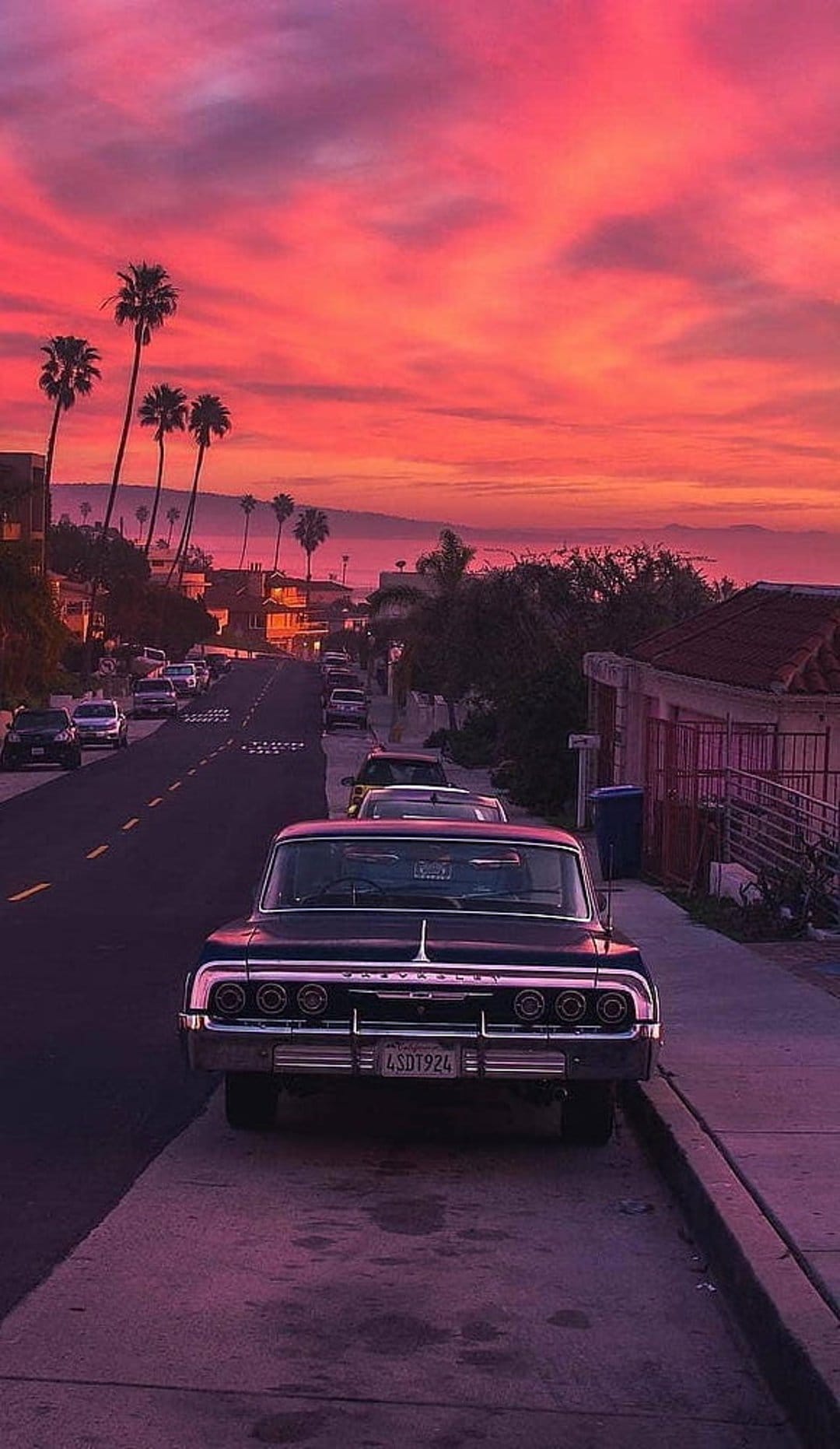 Vintage car parked on a street beneath silhouetted palm trees during a vibrant orange and pink sunset scene.