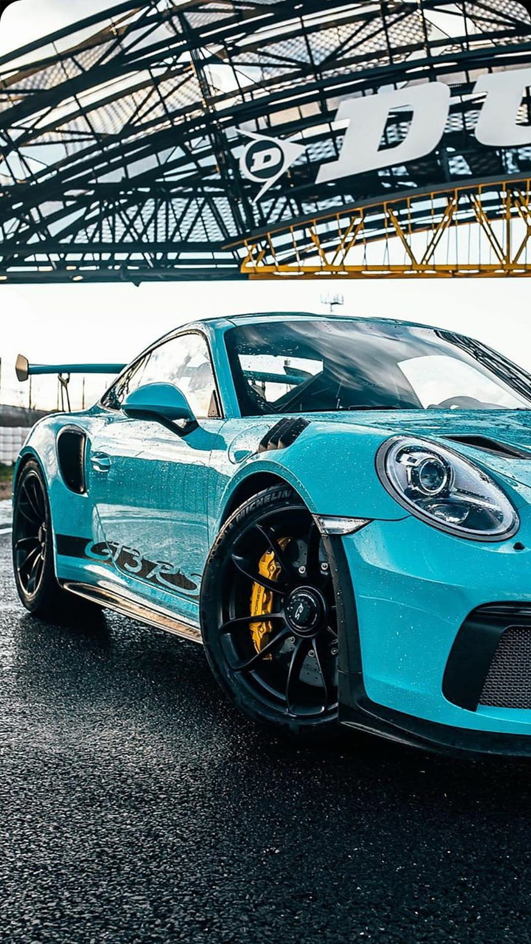 Blue Porsche GT3 RS on a wet asphalt track with yellow brake calipers and the Dunlop bridge in the background.