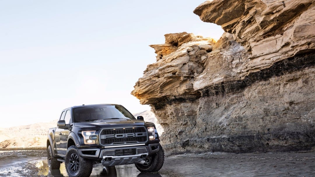 Black Ford Raptor pickup truck parked next to a layered desert cliff under a bright, clear sky background.