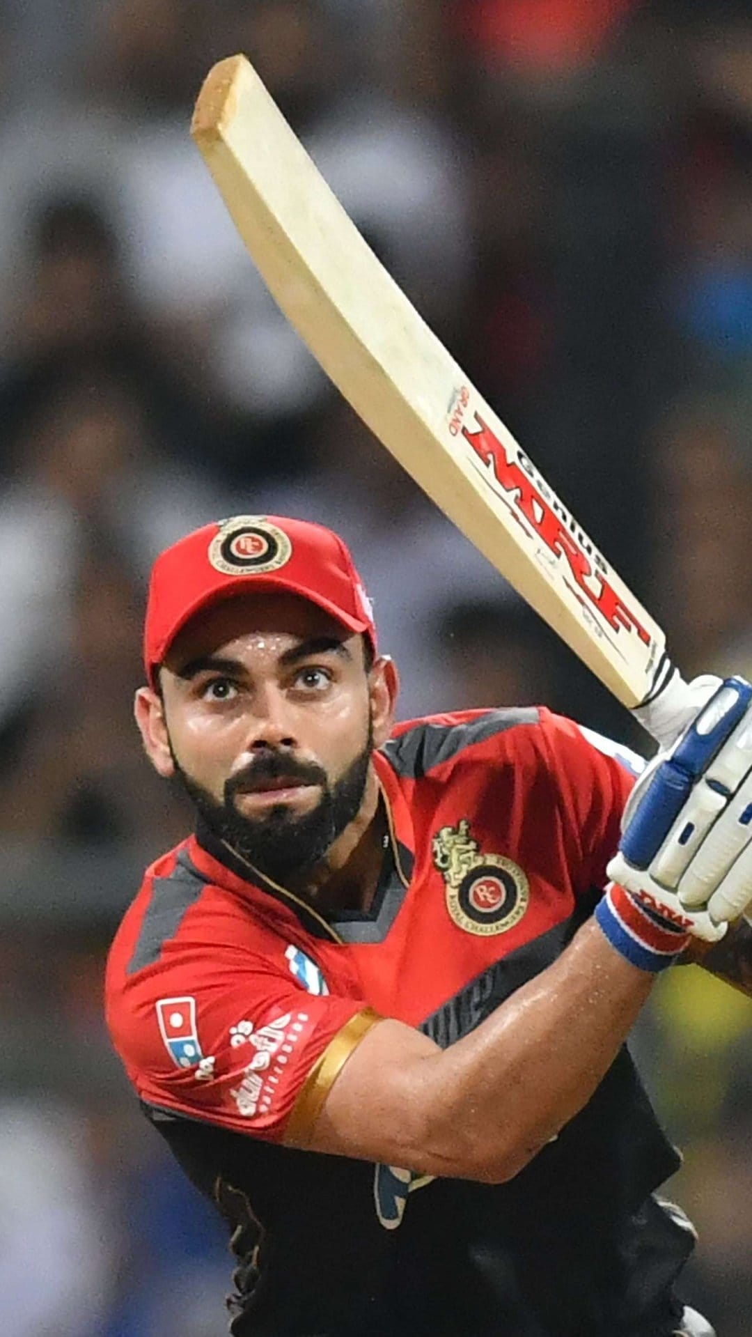Virat Kohli grips his MRF bat while mid-swing wearing a red and black RCB jersey during a cricket match.