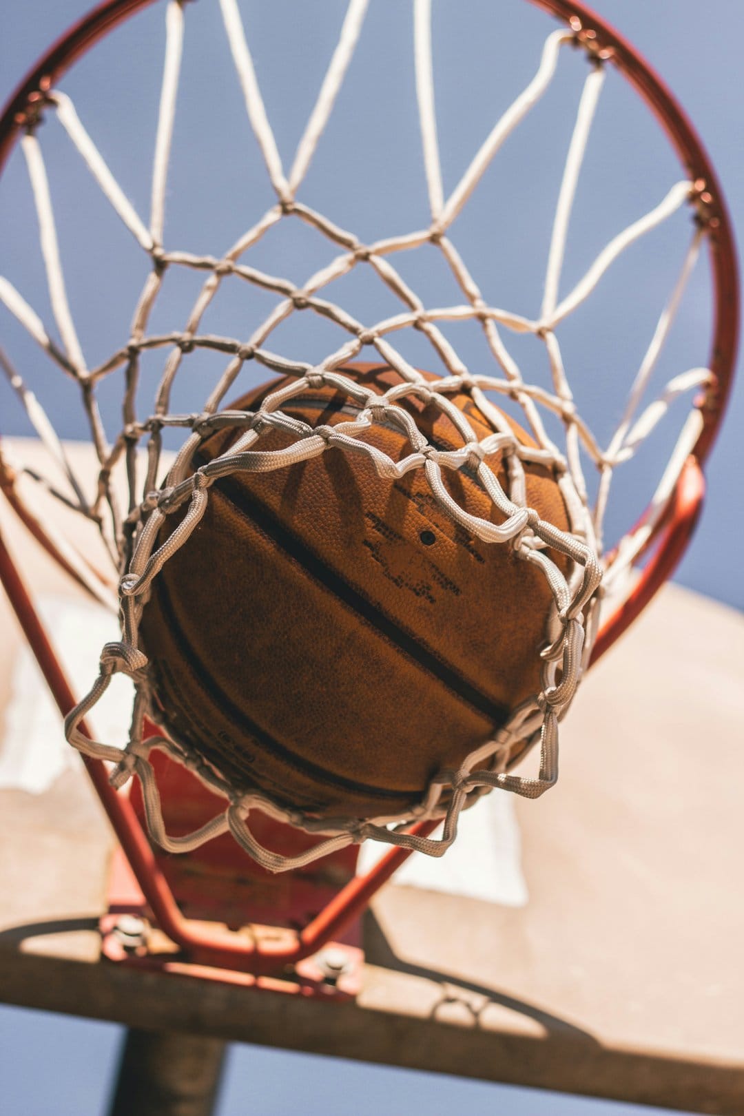 Basketball passing through a white nylon net and red rim against a bright blue sky from a low angle view.