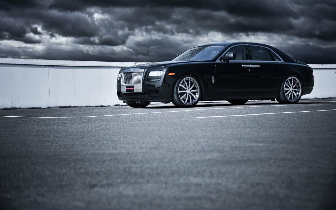 Black luxury sedan with chrome wheels parked on asphalt under a dark stormy sky with a minimalist white wall.