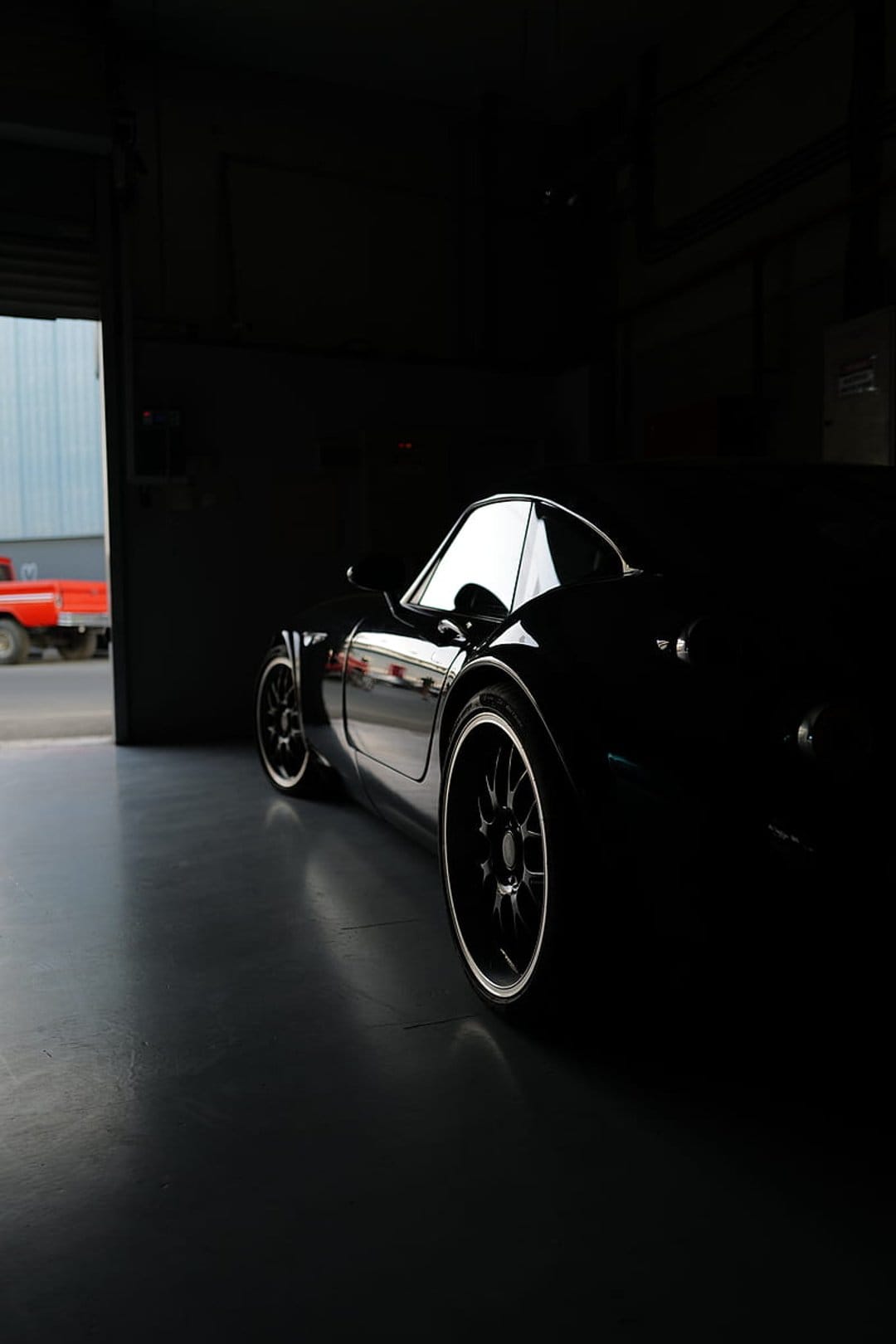Black sports car with glossy rims sits in a dark garage, reflecting dim light across its aerodynamic body panels.