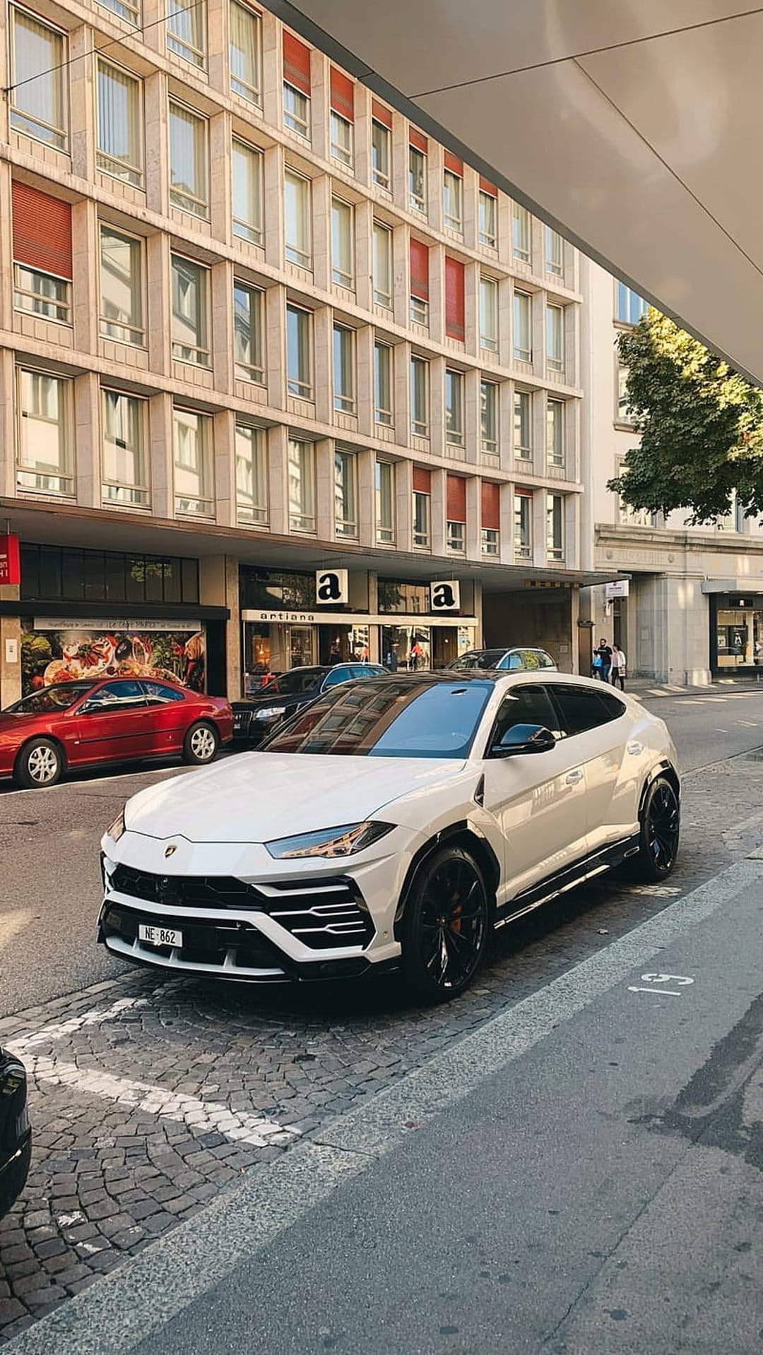 White Lamborghini Urus SUV parked on a gray cobblestone road in front of a modern building with red awnings.