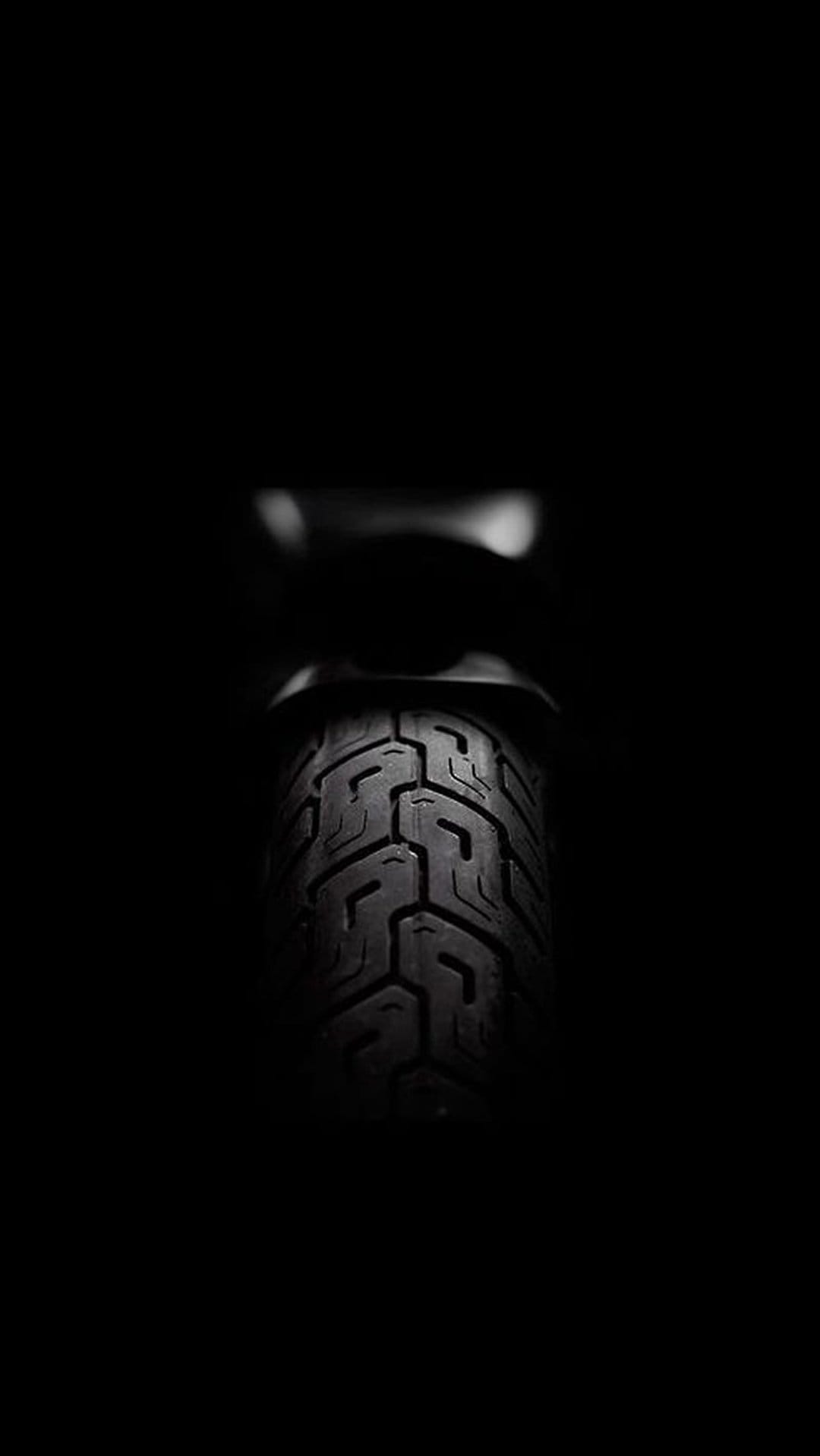 Motorcycle tire tread with deep rubber grooves and sharp high-contrast lighting against a pitch black background.