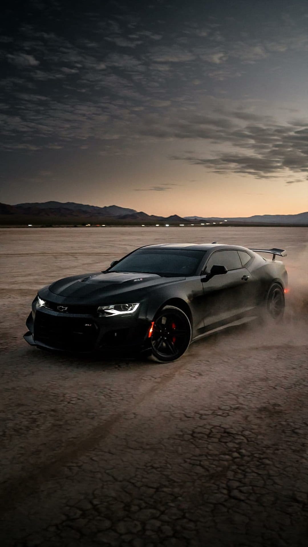 Black muscle car drifts through desert sand during sunset with mountains and heavy dust clouds in the background.