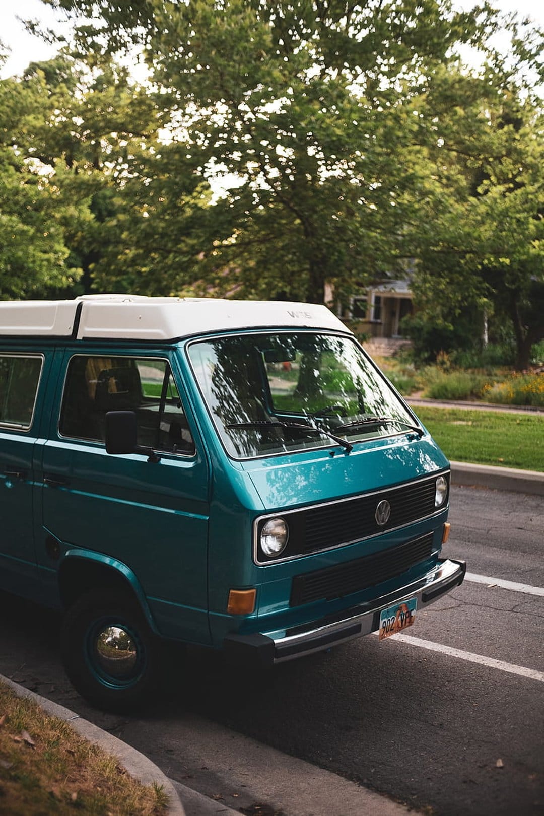 Teal Volkswagen camper van with white pop-top parked on a sun-dappled street under lush green trees.