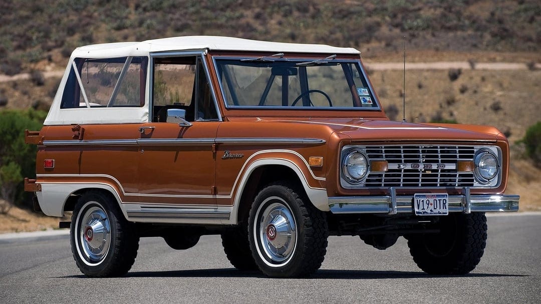 Copper vintage Ford Bronco with white soft top parked on an asphalt road with a blurred desert hill background.