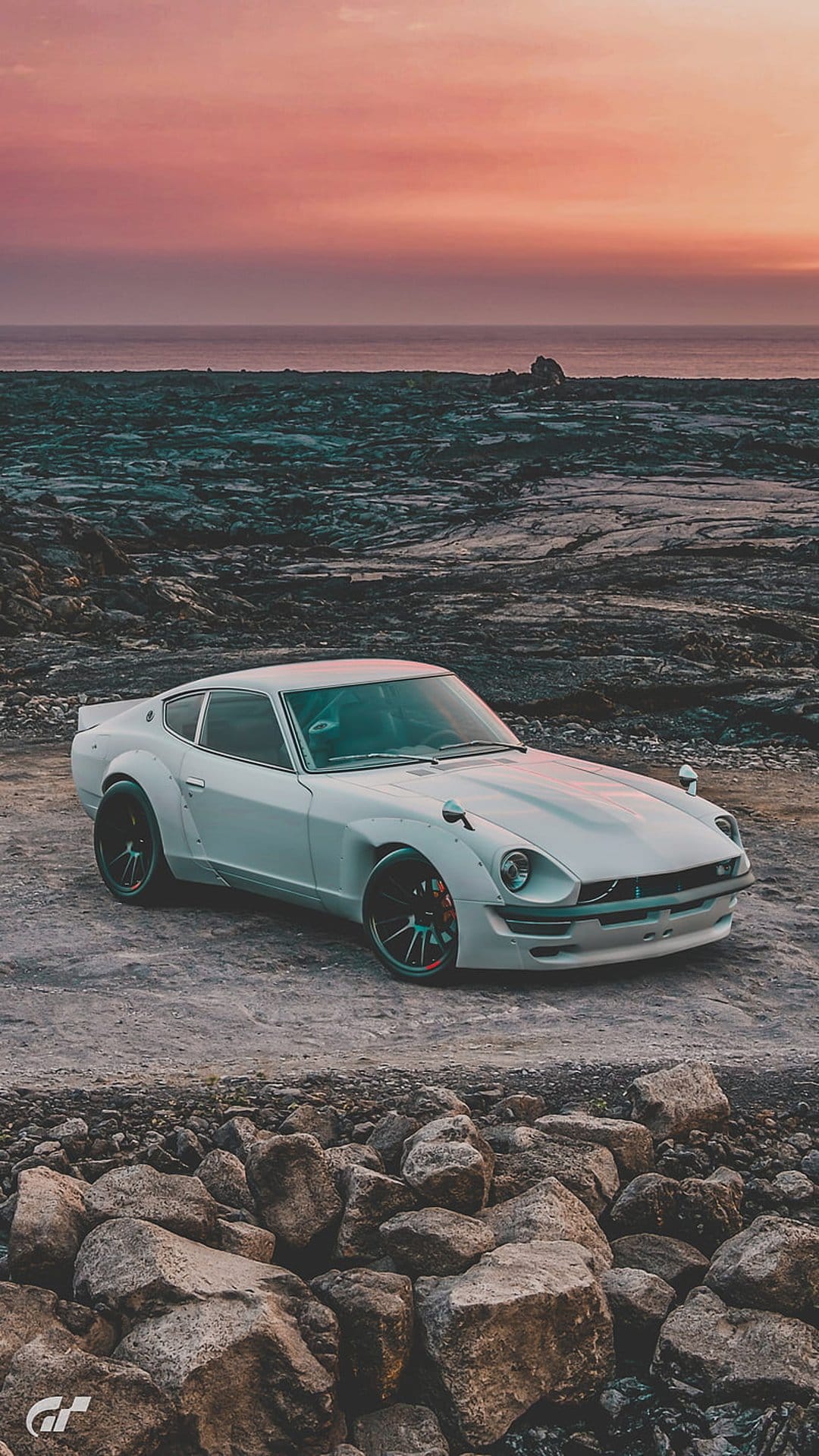 White Datsun Z car on a rocky shore with a vibrant pink and orange sunset sky reflecting over the ocean waves.