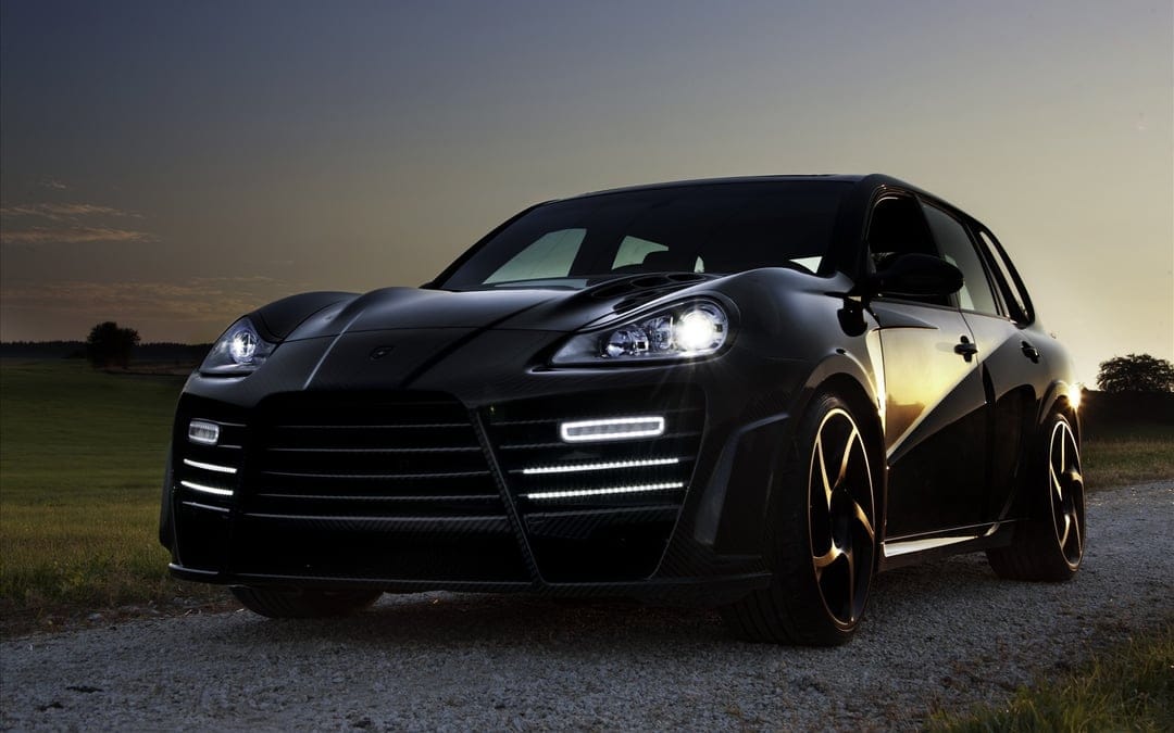 Black modified SUV with bright LED headlights parked on a gravel path under a dark sunset sky.