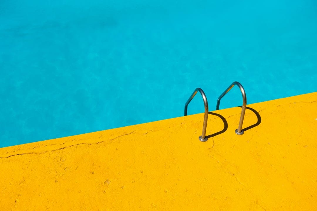 Yellow pool ledge and a metal ladder extending into deep blue water under bright, high-contrast sunlight.