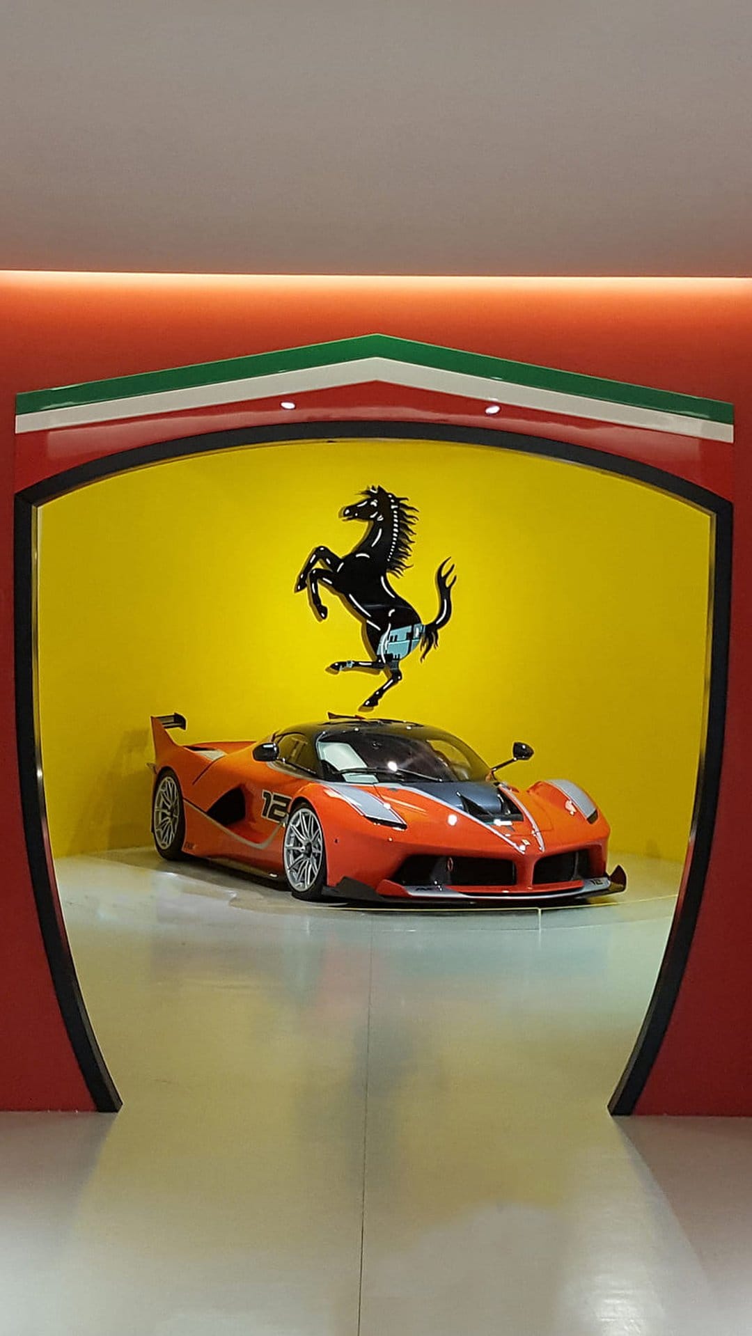 Orange Ferrari FXX K hypercar inside a yellow shield logo featuring the black Prancing Horse and Italian flag.