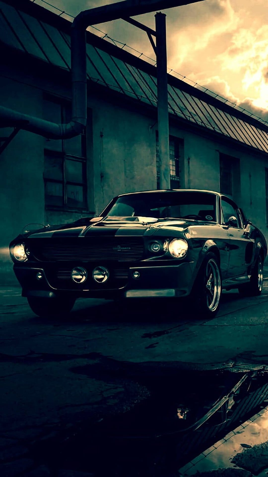 Black Ford Mustang with glowing headlights parked in front of a brick industrial building under a cloudy sky.