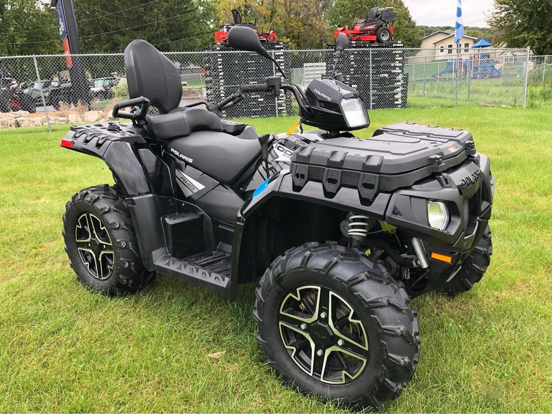 Black Polaris Sportsman Touring ATV parked on green grass with dual seats, chunky tires, and a fence background.